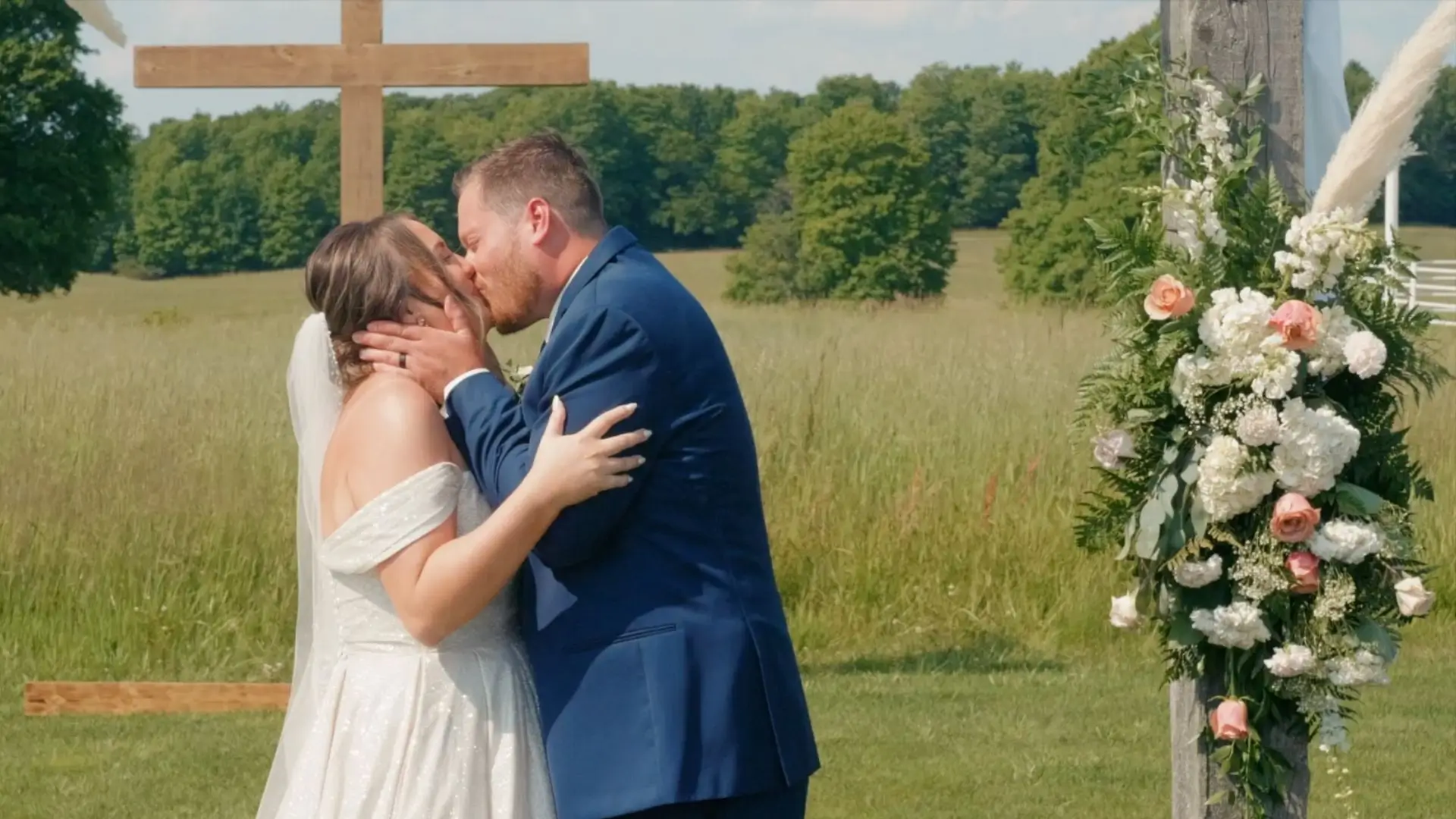 A bride and groom share a kiss at their outdoor wedding at Sonshine Barn in Gaylord. The bride wears a white dress, and the groom is in a blue suit. They are flanked by a wooden cross and floral decorations, with a grassy field in the background.