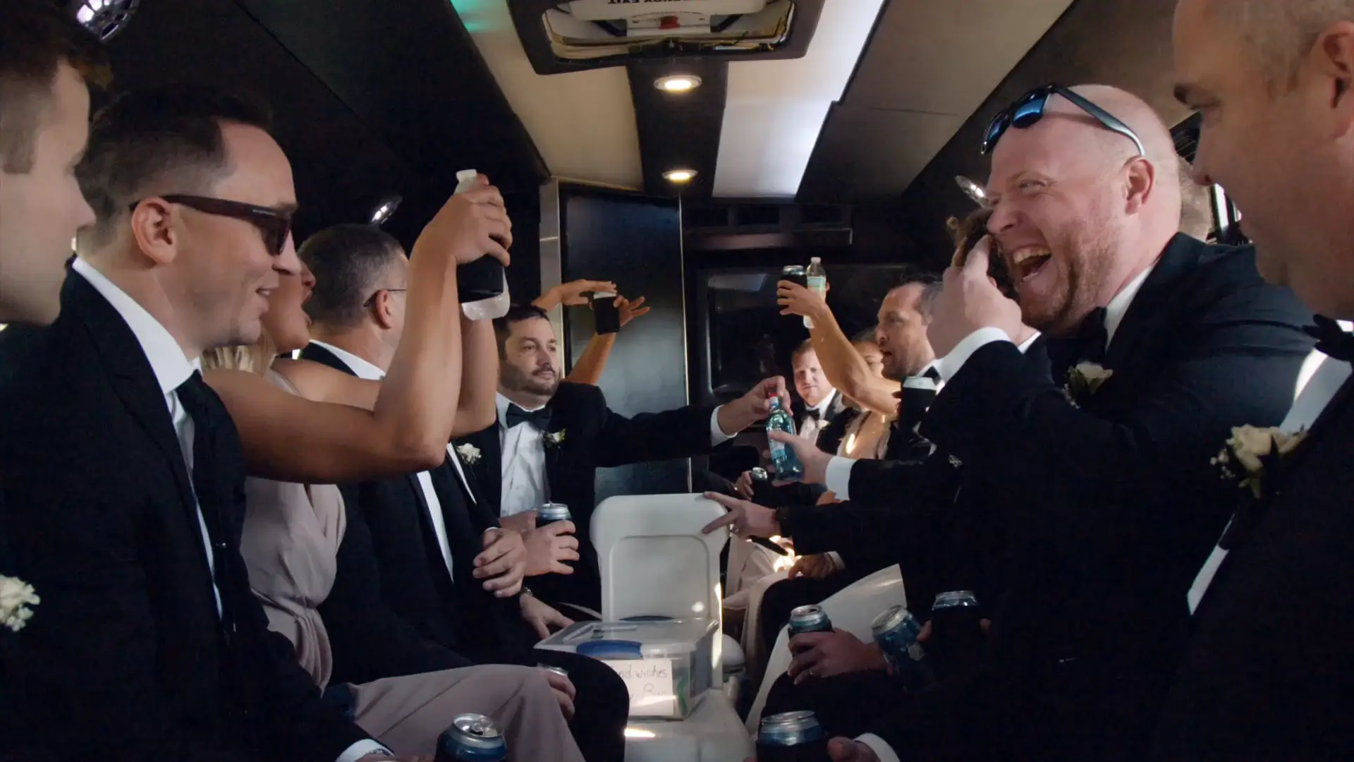 The wedding party celebrates in a lively Michigan limo ride, laughing and raising drinks while wearing formal attire.