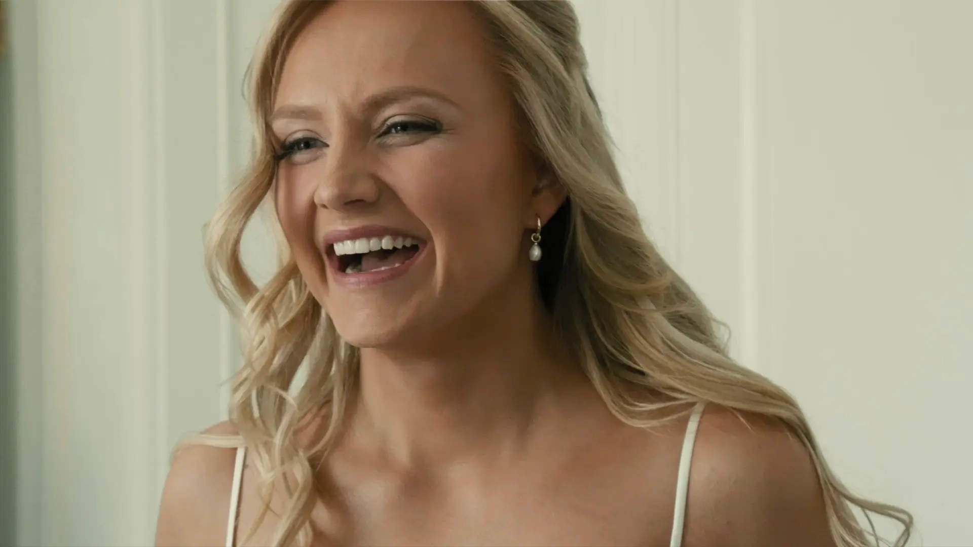 A joyful bride with long blonde hair, wearing pearl earrings and a wedding dress, laughing during her wedding in Michigan.