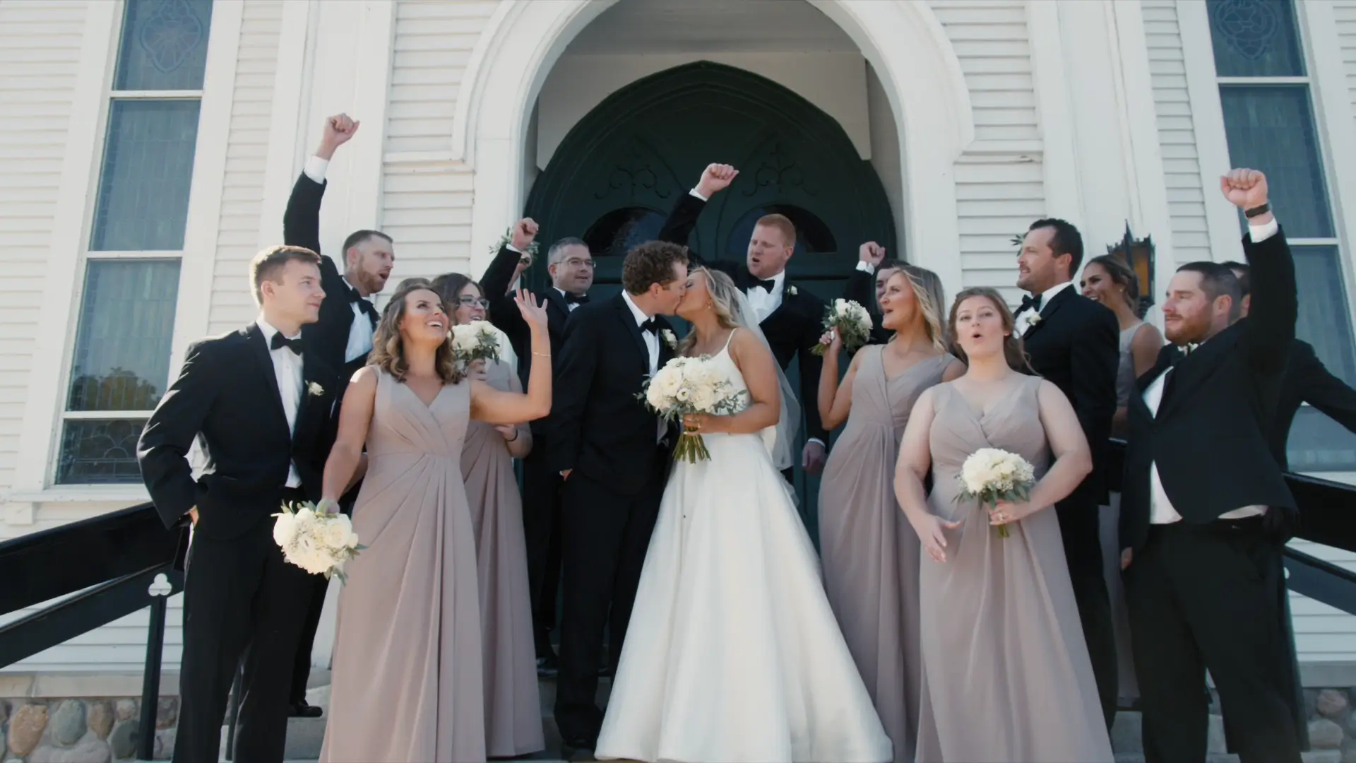 Bride and groom, Kate and Kendrick, kiss outside St. Patrick Parish. Surrounded by a joyful bridal party in elegant attire, they celebrate their Michigan wedding. The group is cheering, with bridesmaids holding bouquets of white flowers.