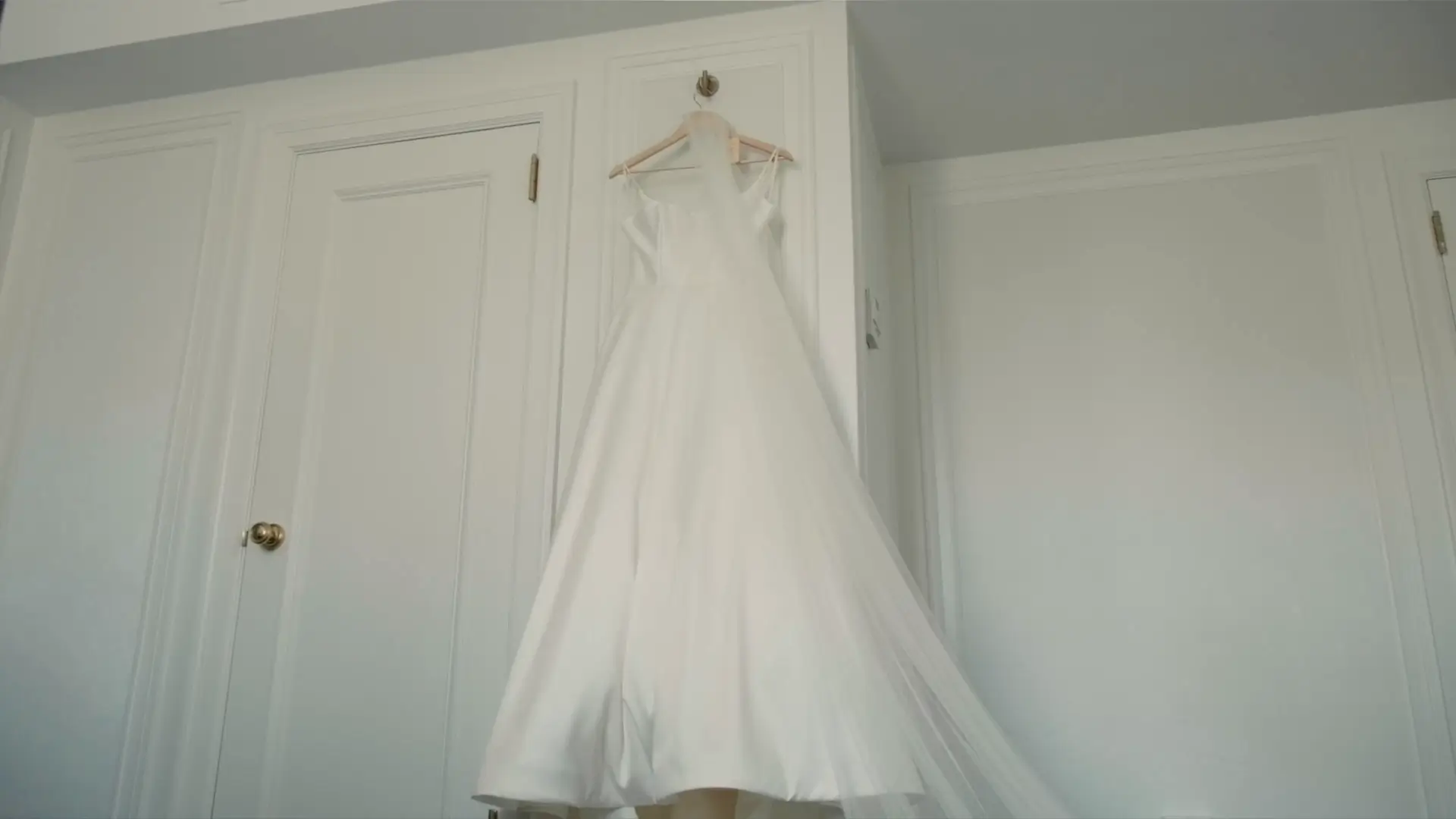 A white wedding dress hangs elegantly from a hook on a white wardrobe door. The dress is full-length with a flowing skirt and sheer veil draping down, set in a bright room, capturing a serene and anticipatory moment before Kate and Kendrick's wedding.