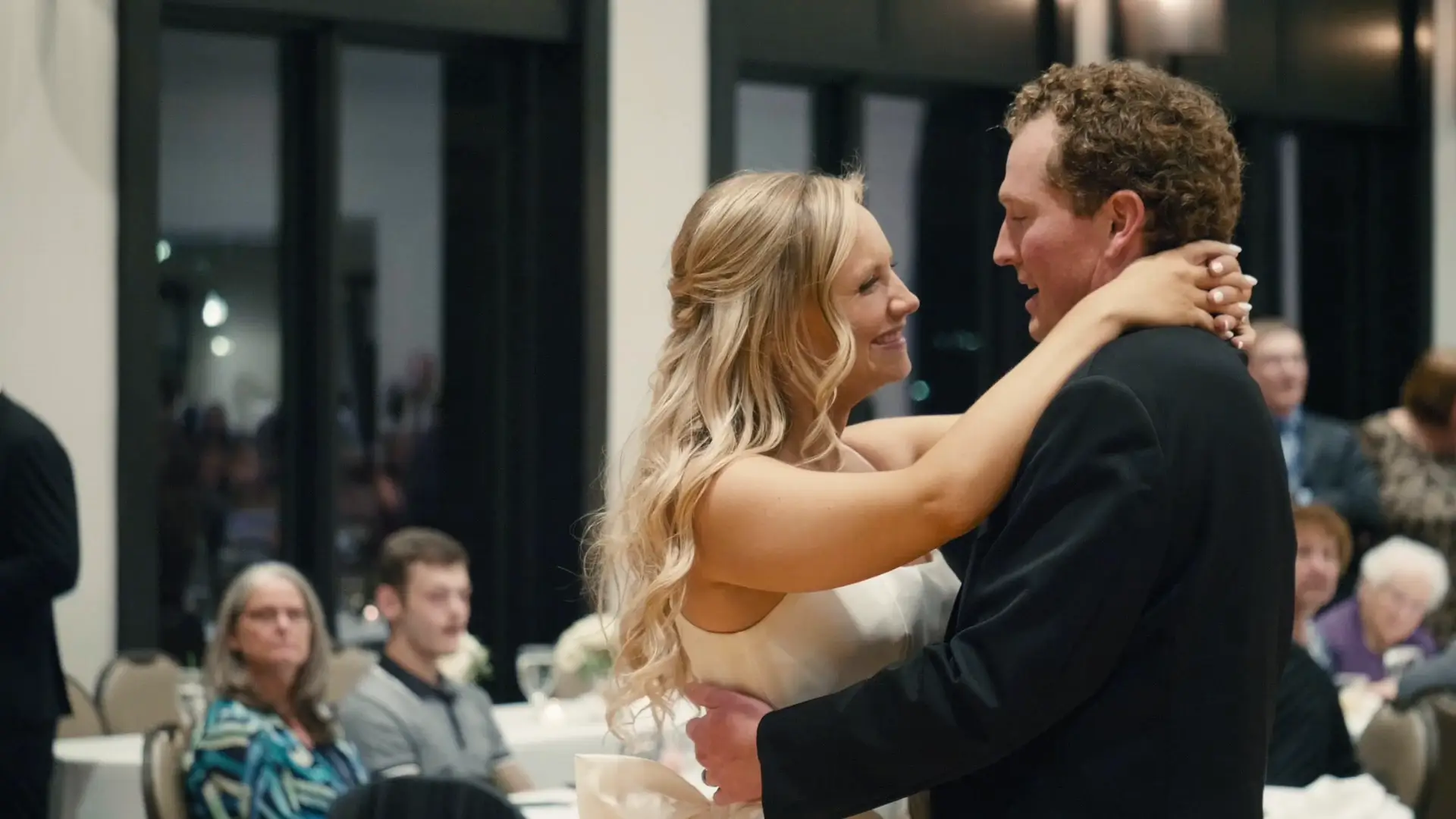 Kate and Kendrick share a first dance at their wedding reception. Kate is in a white dress, smiling at Kendrick, who is in a dark suit. Guests can be seen sitting at tables in the background, watching the couple.