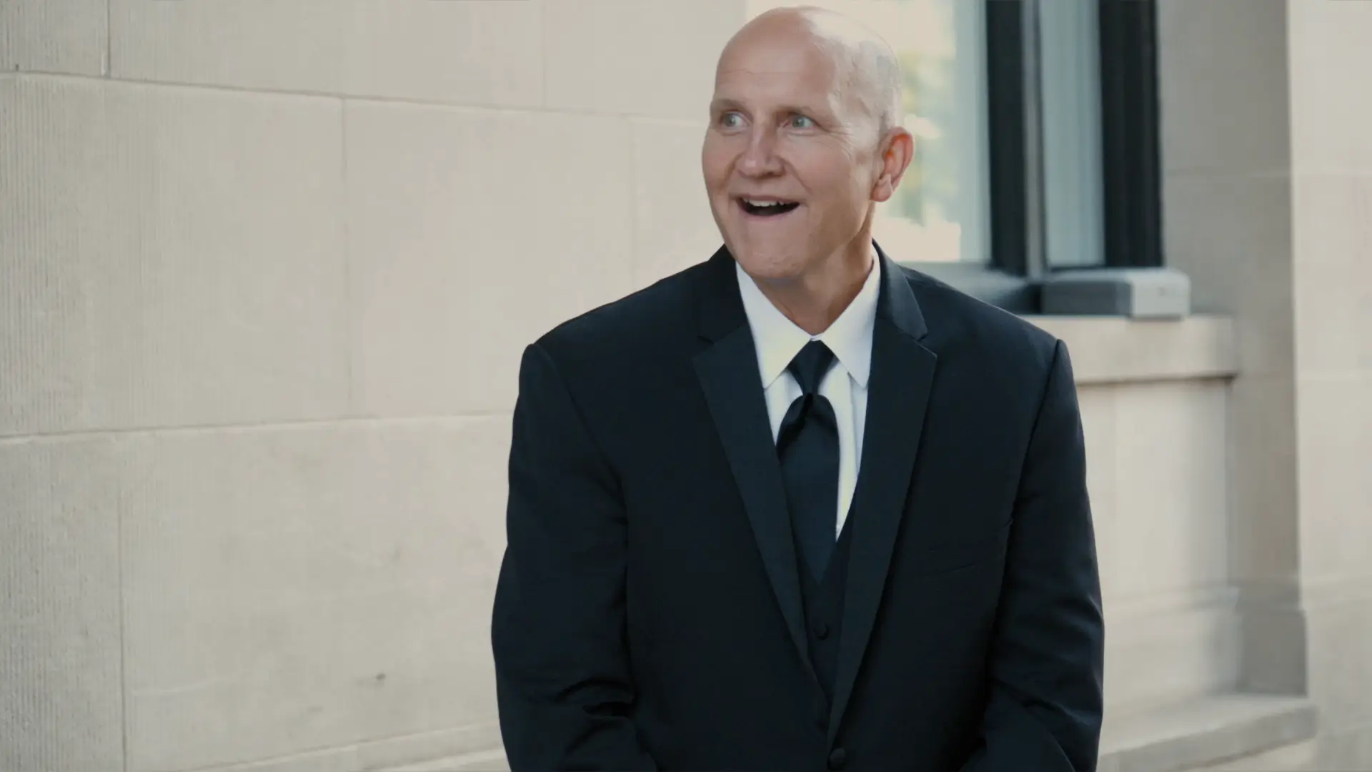 A man in a tuxedo stands against a stone wall, looking to the side with a joyful expression. Sunlight gently illuminates the scene, capturing a candid, happy moment at a Michigan wedding.
