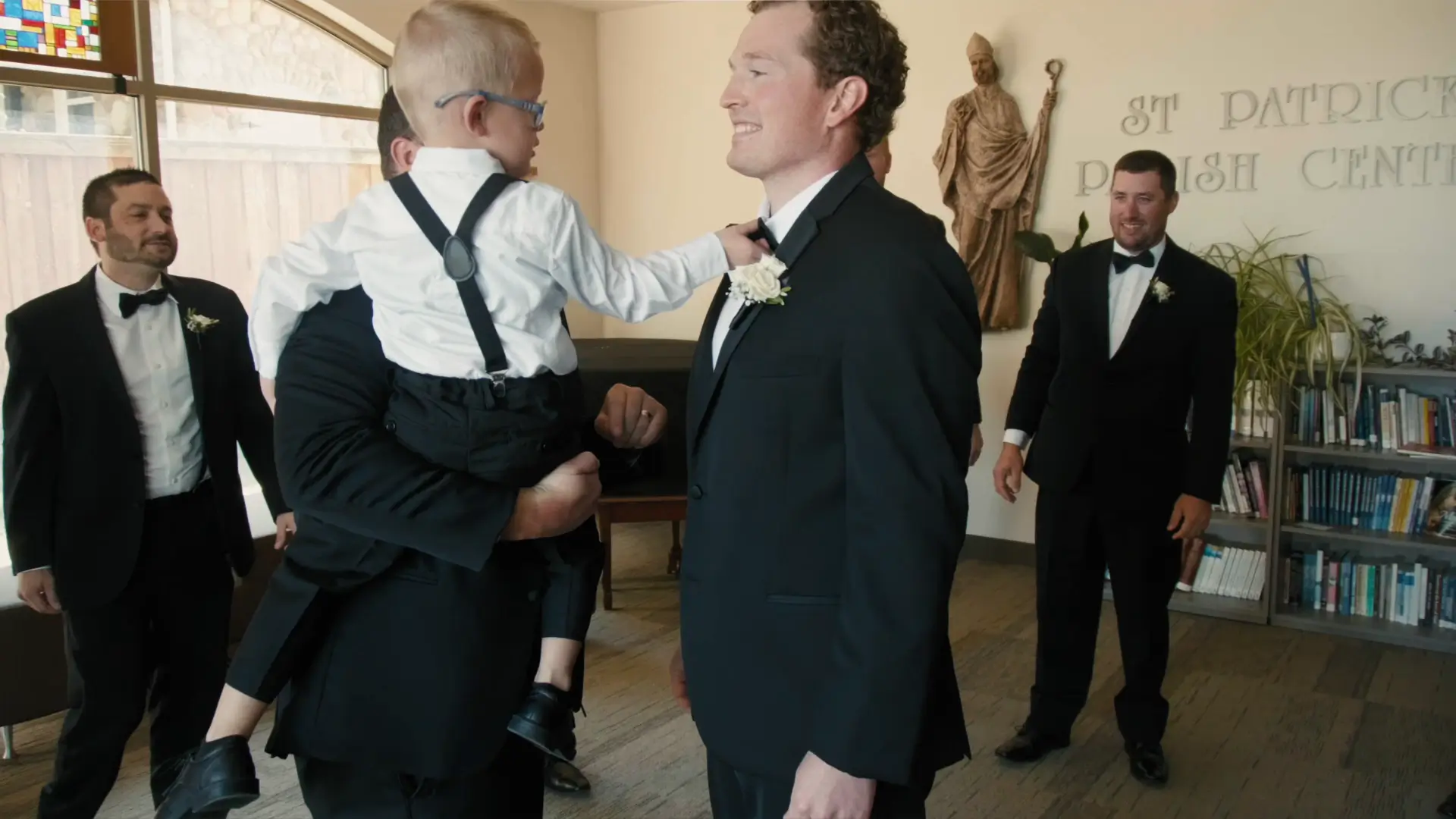 Groom in a tuxedo smiles warmly at a child dressed similarly, held by a groomsman. Two other groomsmen stand nearby, all in formal attire, inside St. Patrick Parish Center with a statue and bookshelves visible in the background.