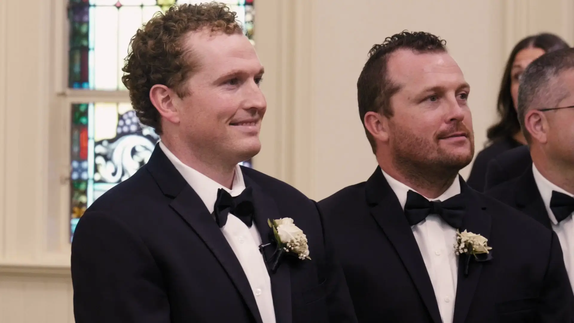 Groomsmen in black tuxedos with boutonnieres, smiling inside St. Patrick Parish in Grand Rapids, Michigan. A stained glass window is in the background.