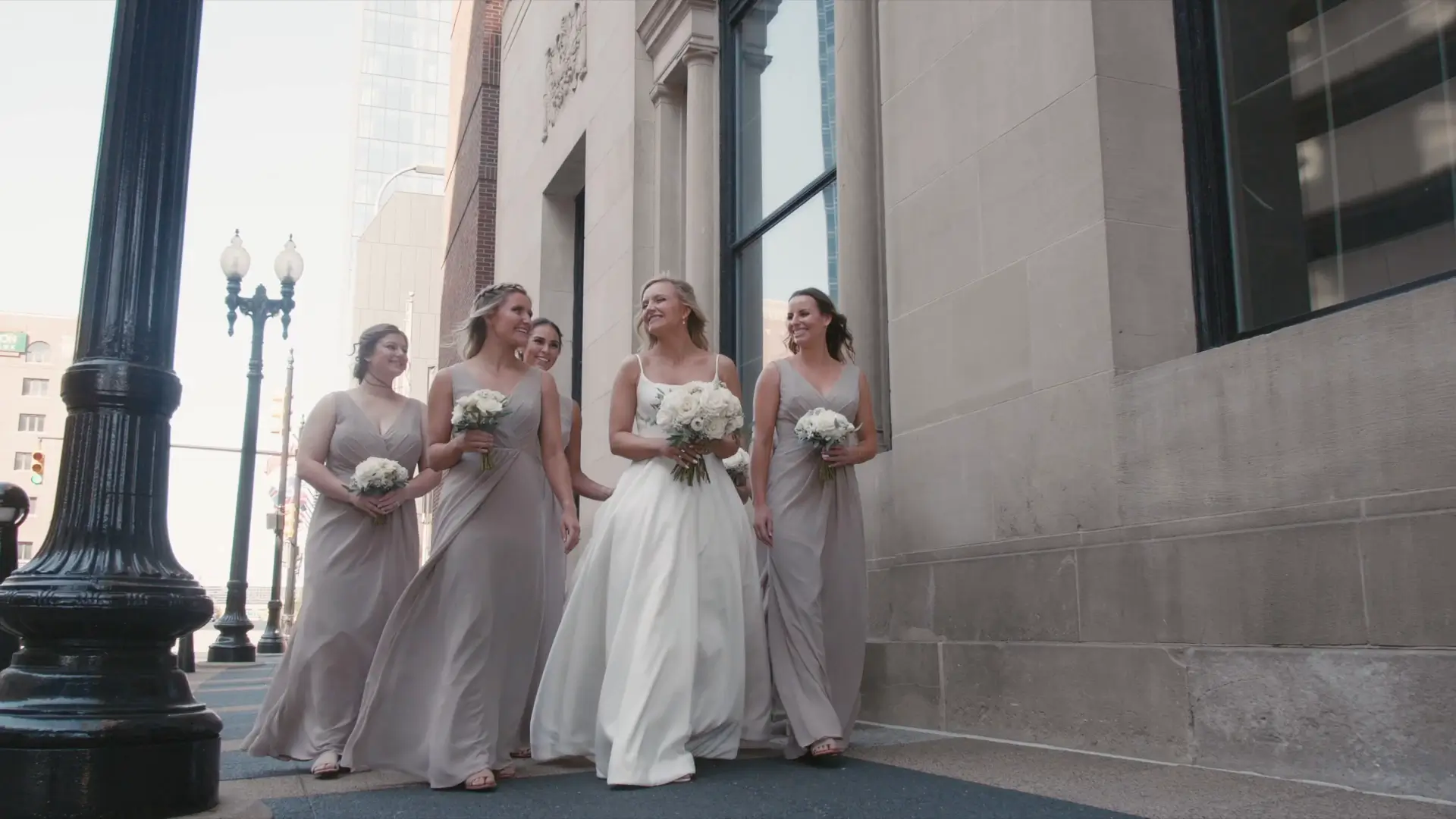 Bride in a white gown walks joyfully with her bridesmaids, holding bouquets of white flowers. They are on a city sidewalk next to a building with large windows, under a streetlamp. The scene depicts a cheerful pre-wedding moment.