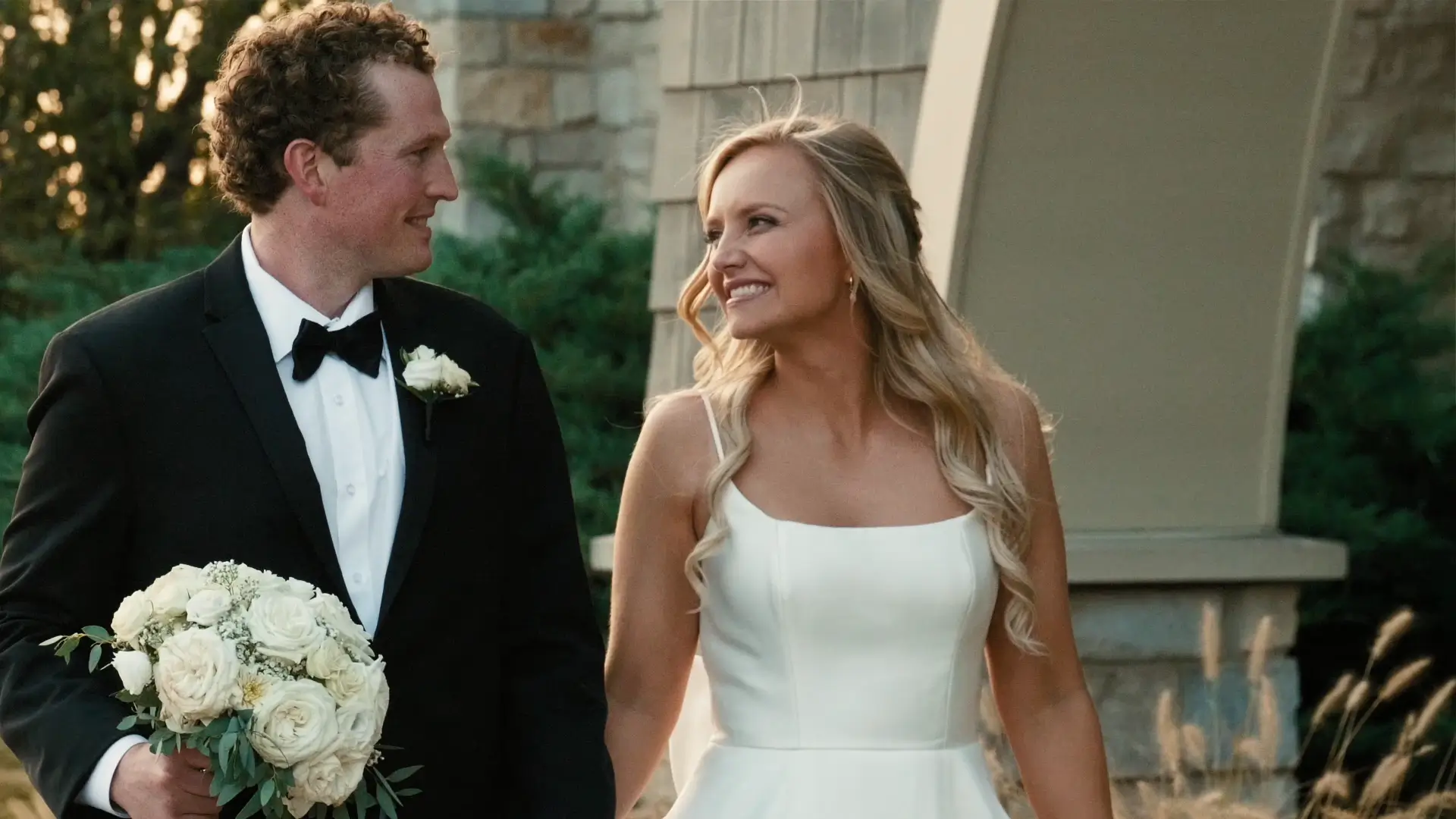 A groom in a black suit holds a bouquet of white roses, walking hand in hand with the bride in a white dress. They smile at each other against a backdrop of stone architecture and greenery.