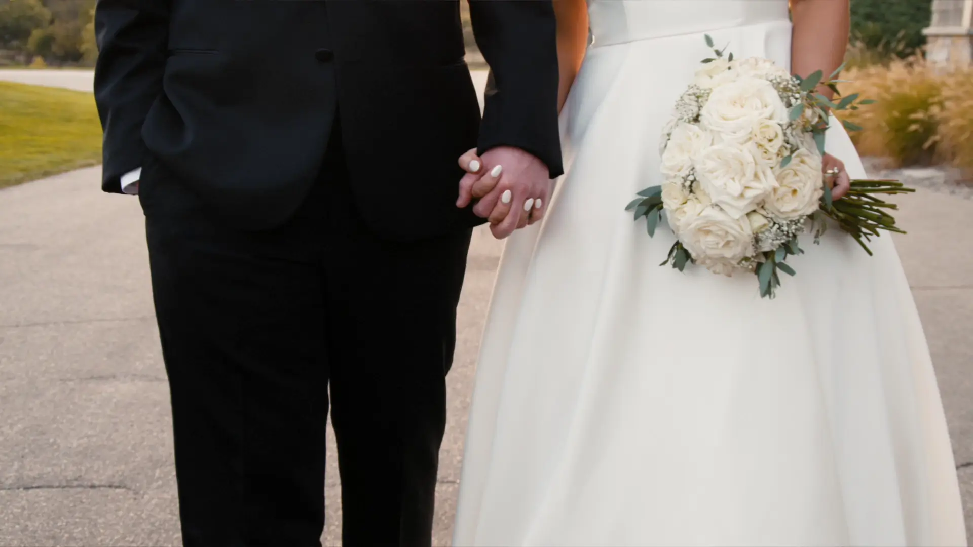 Bride and groom holding hands. Bride in a white dress holding a bouquet of white roses, groom in a black suit. Outdoors on a path with grass in the background.