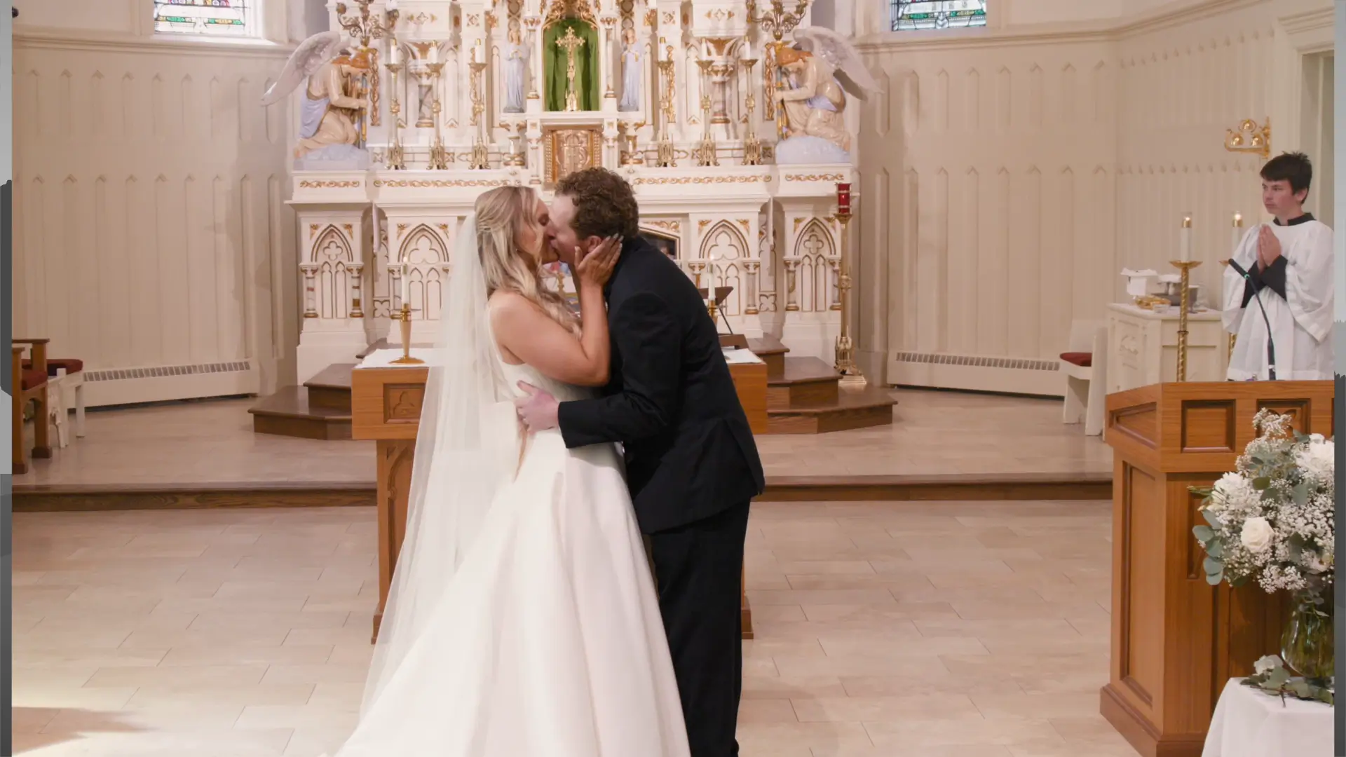 A bride and groom share a kiss at the altar inside St. Patrick Parish in Ada, Michigan, during their wedding ceremony. The ornate altar is visible in the background, and an altar server stands to the right.