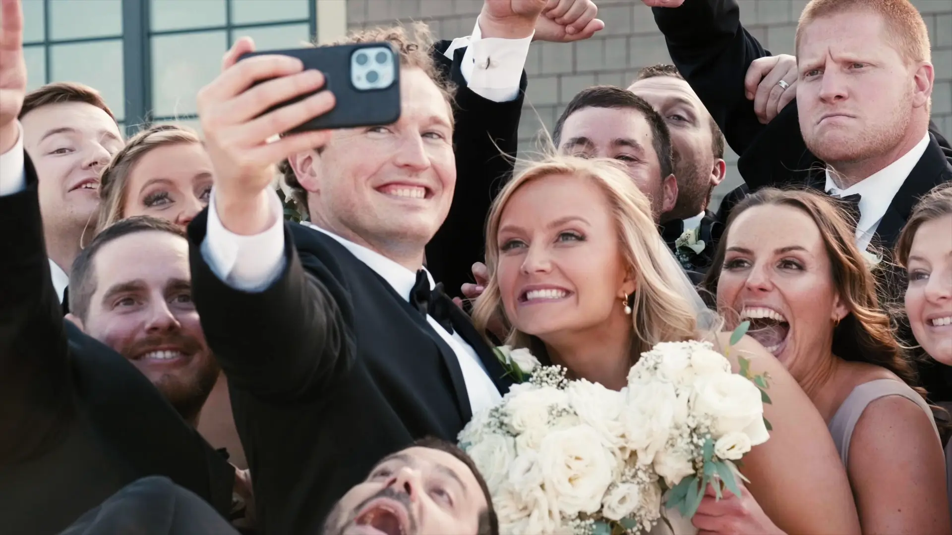 The bride, groom, and their wedding party taking a joyful selfie. The bride holds a white bouquet, and everyone is smiling and cheering. They are dressed in formal attire, capturing a candid, celebratory moment outside.