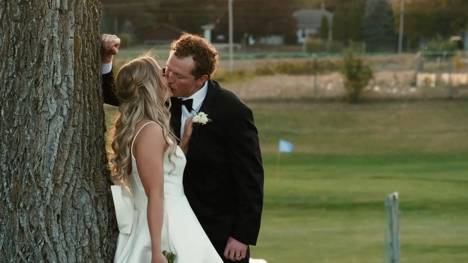 Bride and groom share a kiss by a tree, set against lush Michigan greenery. Bride in a white dress with a delicate bow and groom in a black tuxedo with a boutonniere. The backdrop includes serene fields, capturing a romantic wedding moment.