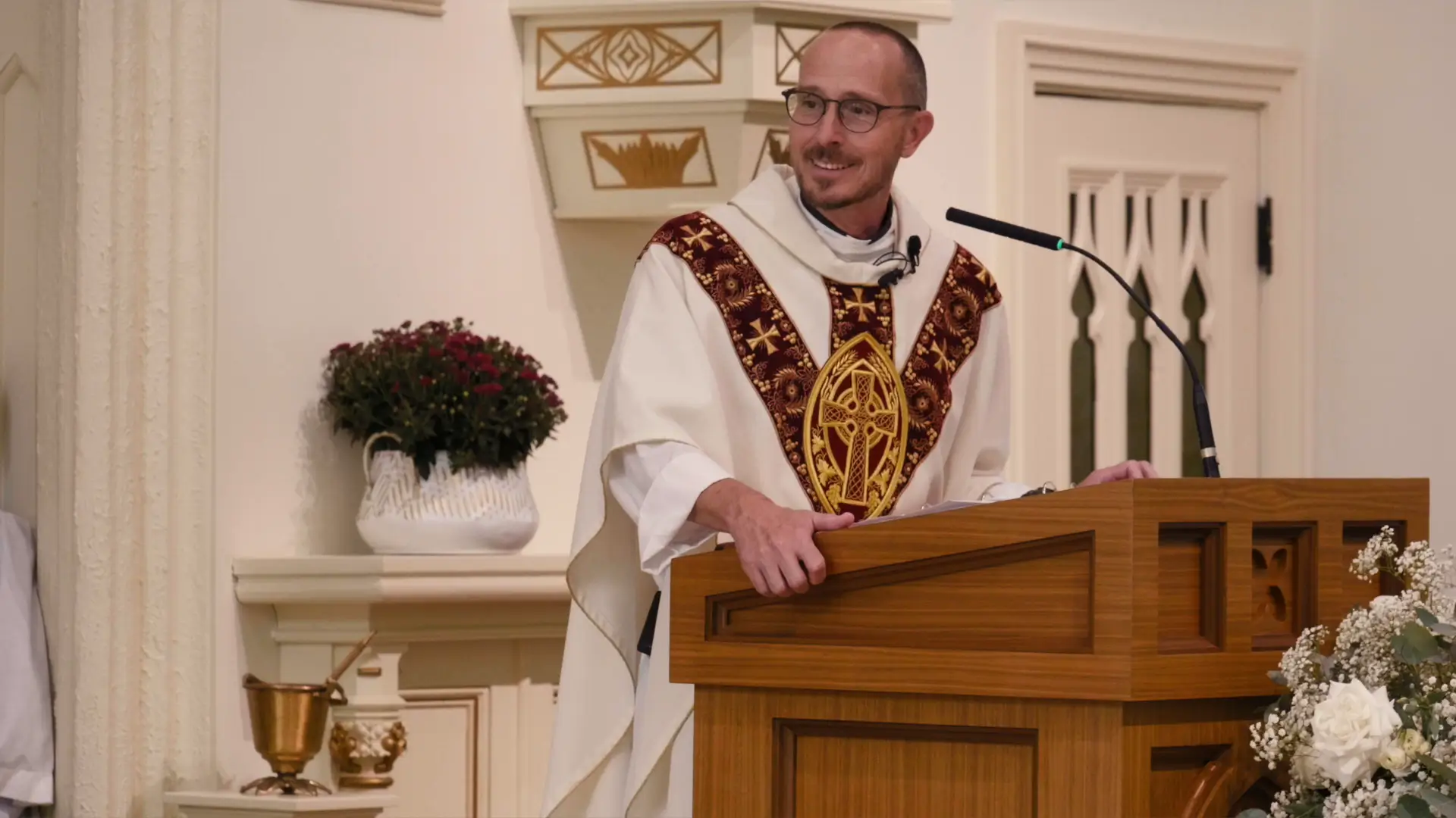 Priest standing at a wooden lectern, smiling, during a ceremony at St. Patrick Parish, Ada, Michigan. The priest wears ornate robes and glasses. A vase with flowers and a ceremonial brass vessel are visible nearby.