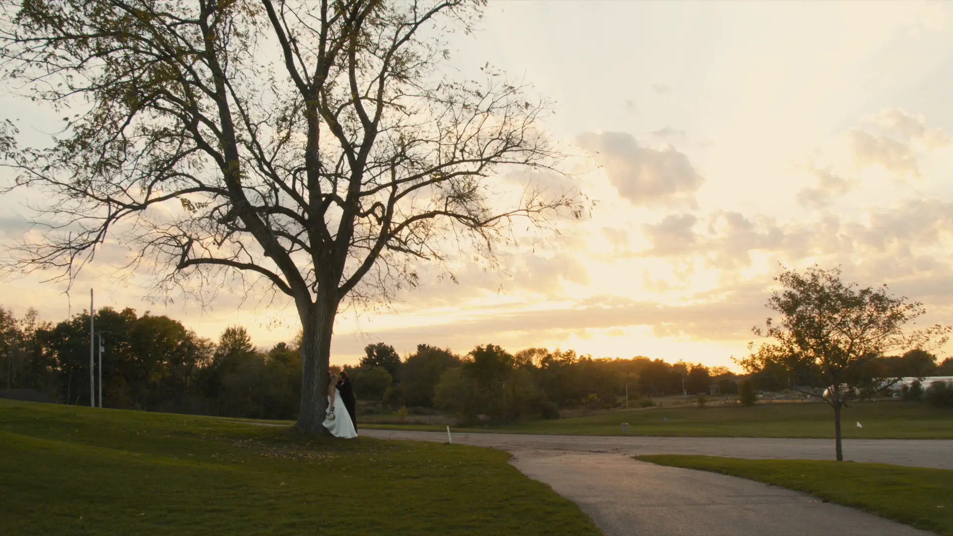 Bride and groom standing under a large tree at sunset, with a pathway and grassy landscape in the foreground. The sky has a warm, golden glow with scattered clouds.