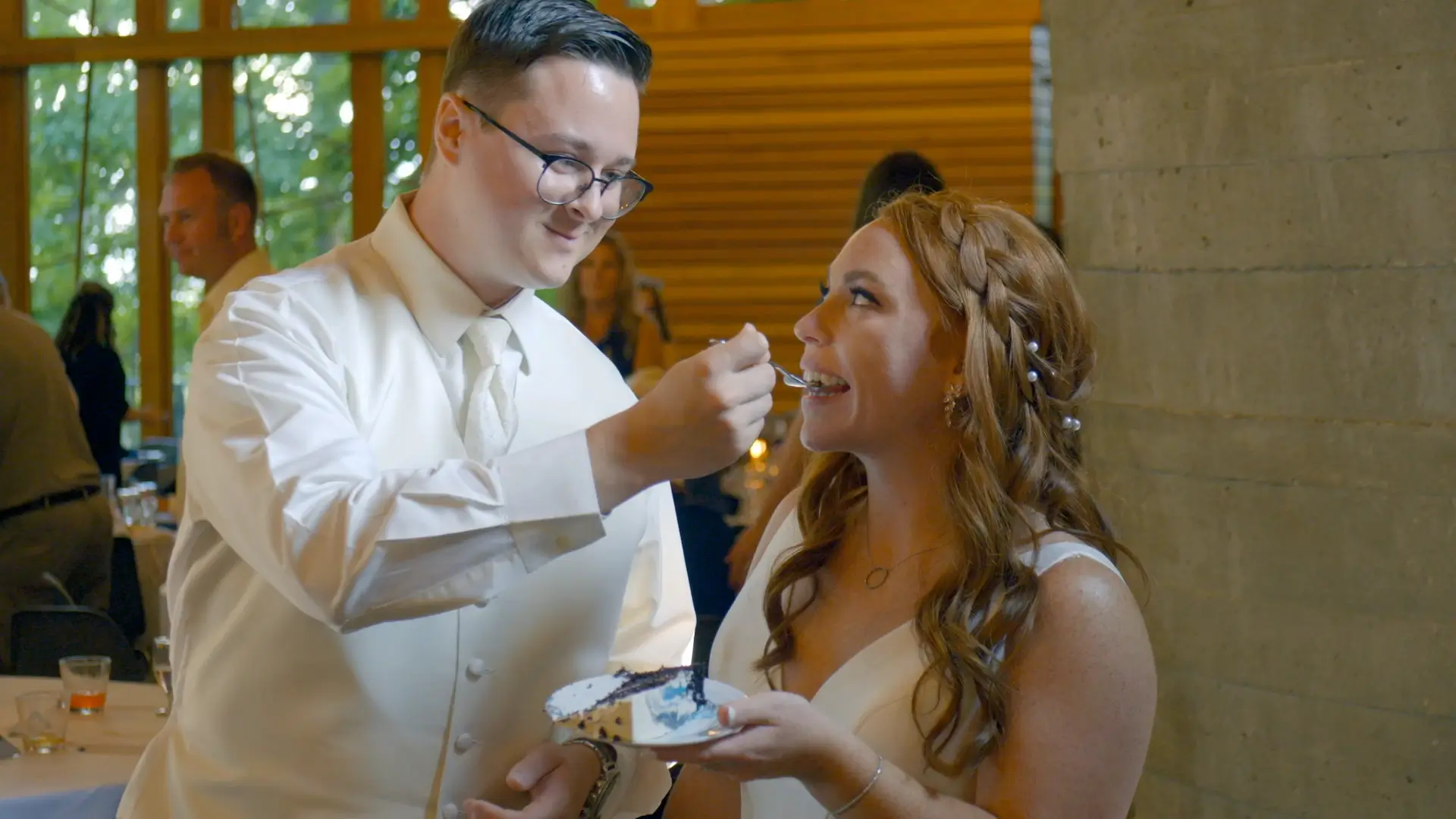The groom, dressed in a white vest and tie, smiles as he feeds a piece of cake to the bride, who holds a plate and looks up at him with a smile during their wedding reception at the Bissell Tree House in Grand Rapids. Guests are visible in the background.