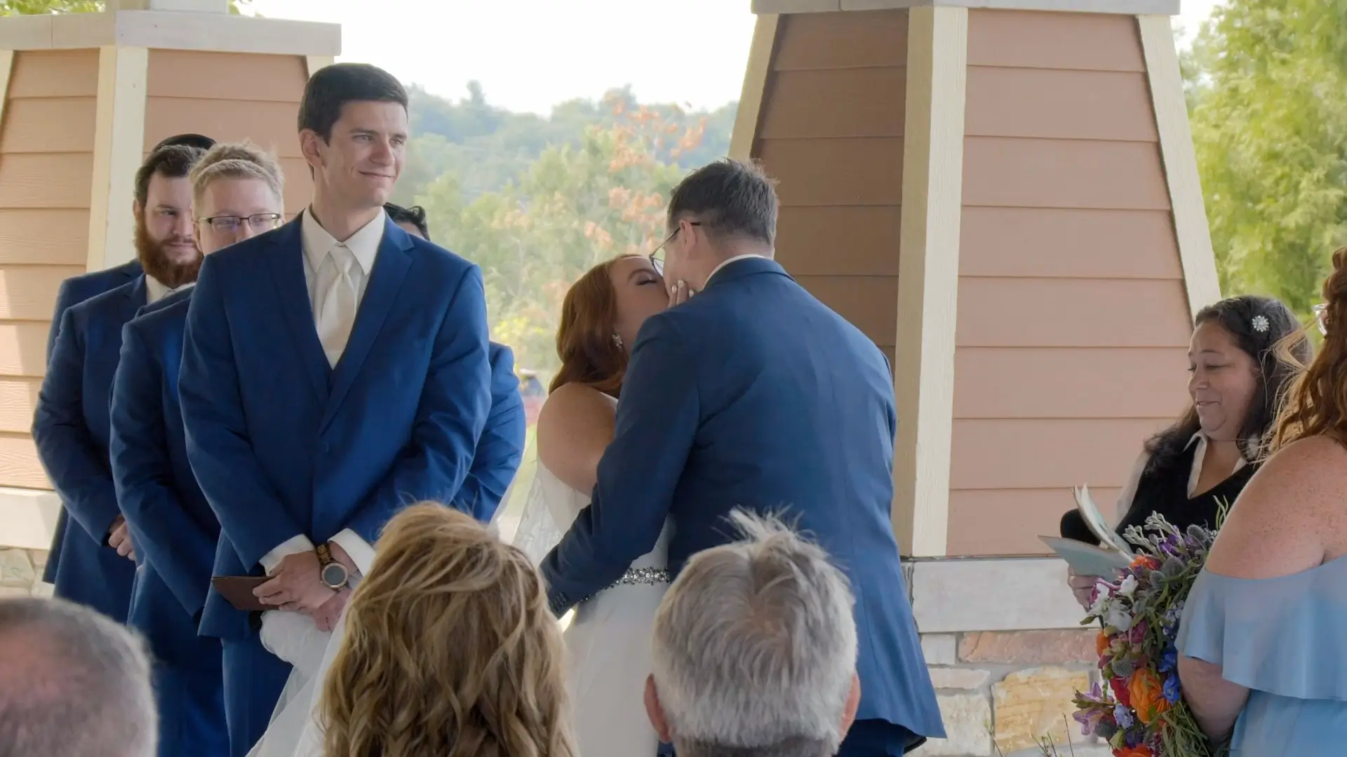 Sydney and Jacob share their first kiss at the altar during an outdoor wedding ceremony at The Bissell Tree House at the zoo. The bridal party and guests smile on a warm summer day.
