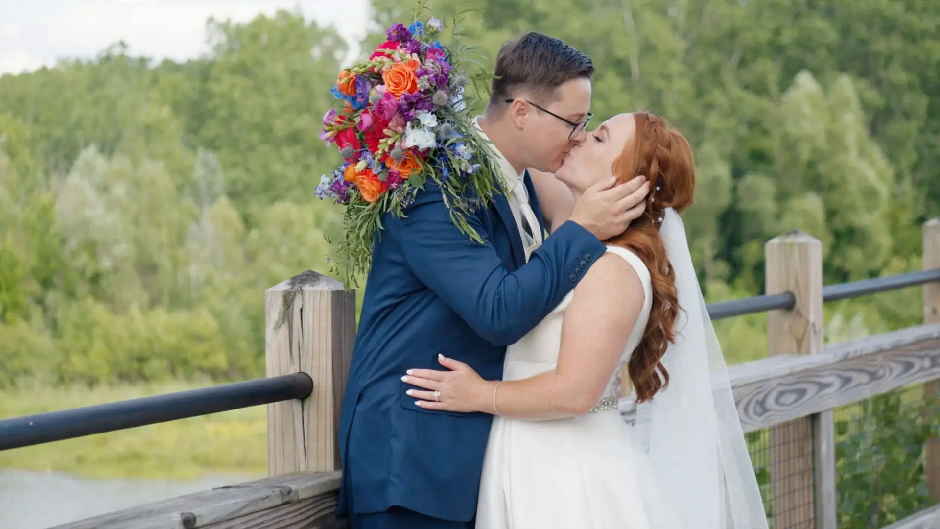 A couple, Sydney and Jacob, share a kiss surrounded by lush greenery at the Bissell Tree House in Grand Rapids. Sydney wears a white dress, and Jacob dons a blue suit while holding a vibrant bouquet. The nature-themed wedding celebration radiates joy.