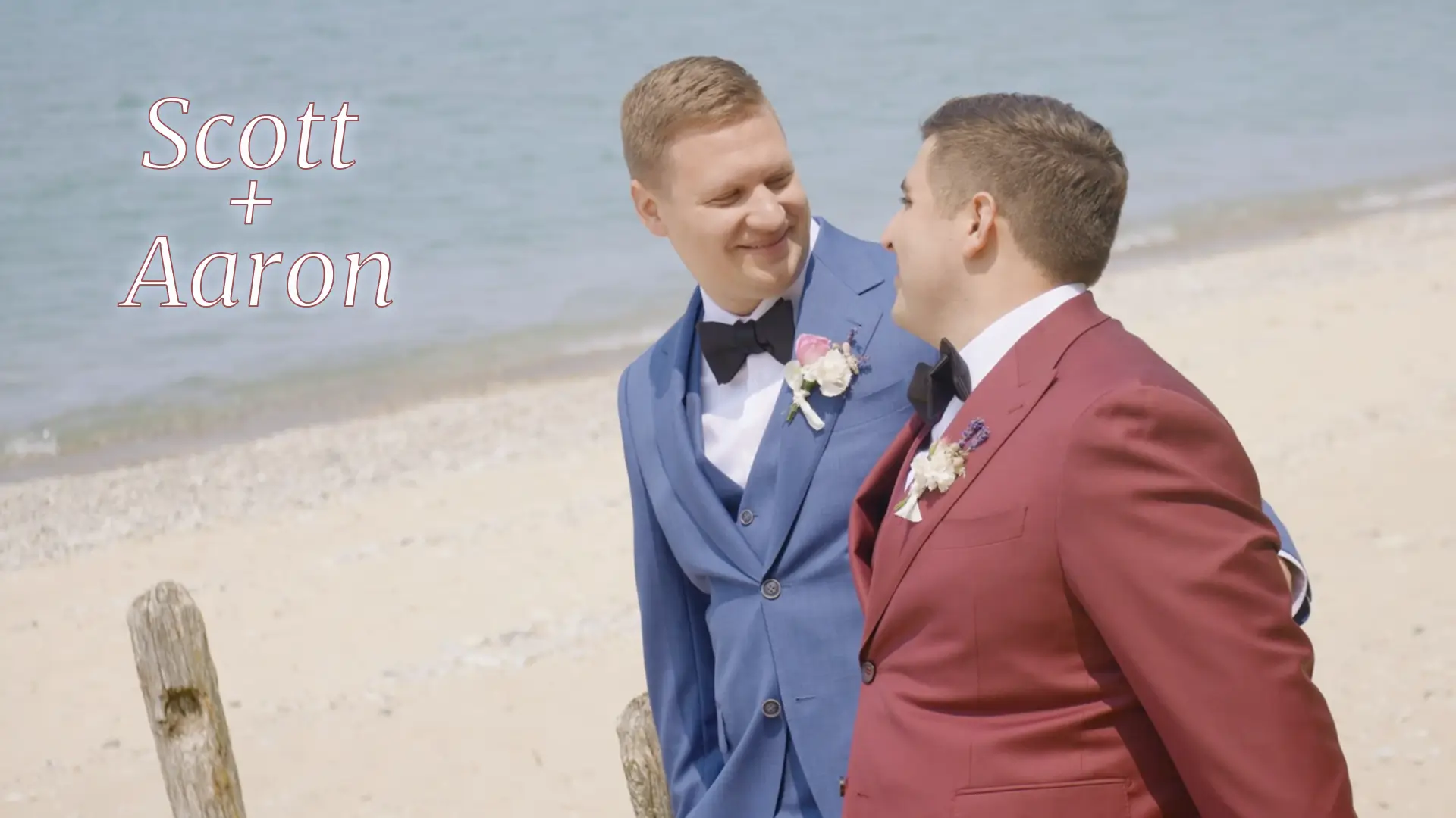 Scott and Aaron, dressed in a blue suit and a red suit respectively, smile at each other while walking on a beach with Lake Michigan in the background. Text overlay reads “Scott + Aaron” in an elegant font.