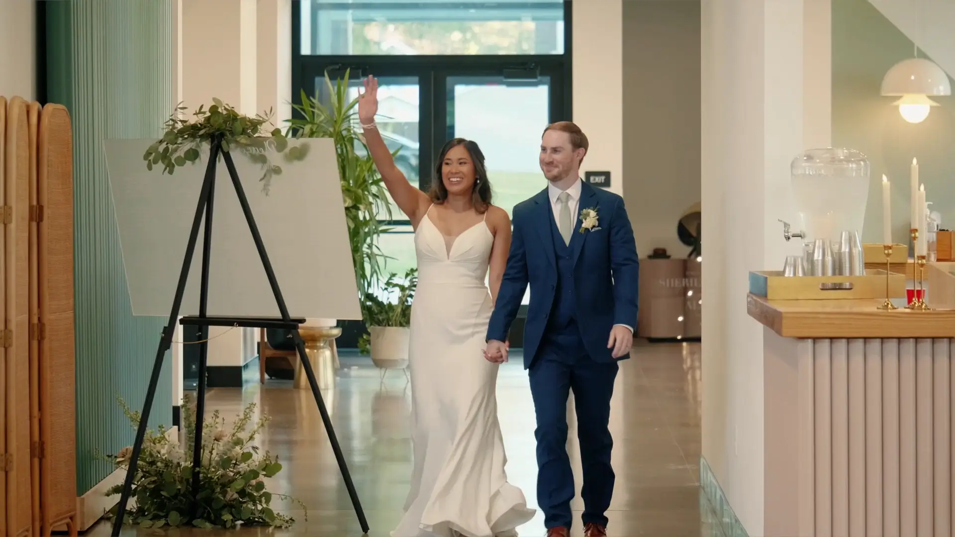 Sherilyn waves joyfully as she and Alex walk hand-in-hand down a hallway at their wedding venue, Leona Road. Captured in this snapshot from their wedding highlight video, the couple exudes happiness on their special day in Grand Rapids, Michigan.