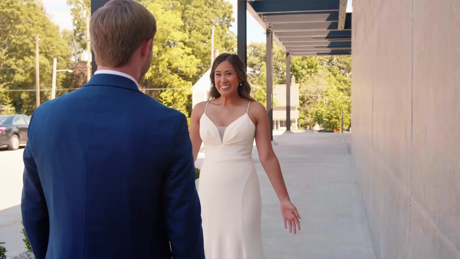 A bride in a white dress smiles at a groom in a blue suit during an outdoor first-look moment. They stand on a sidewalk near a building with a backdrop of green trees under a sunny sky in Grand Rapids, Michigan.