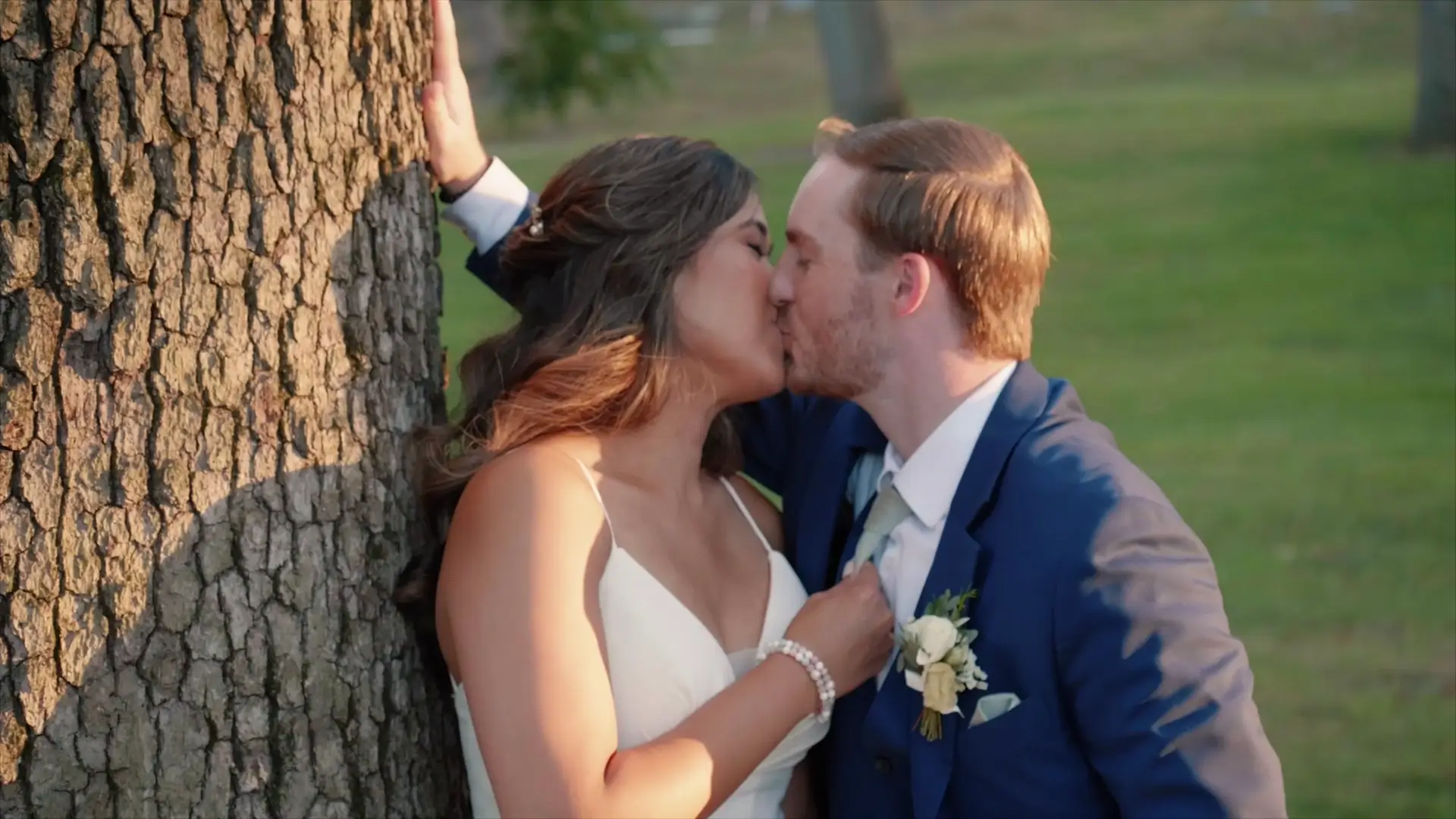 Sherilyn and Alex share a romantic kiss leaning against a tree at Richmond Park in Grand Rapids, Michigan. Sherilyn is wearing a white dress, and Alex is in a blue suit with a boutonniere. The sun casts a warm glow, creating a serene backdrop.