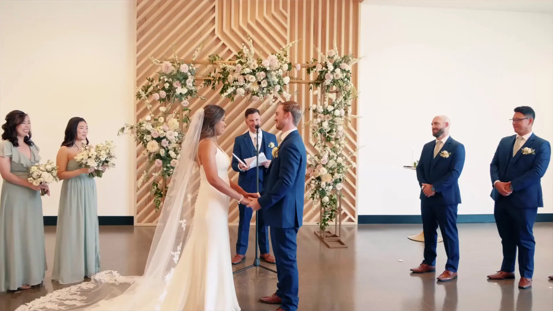 In this snapshot from their wedding video at Leona Road, Grand Rapids, Sherilyn and Alex exchange vows amidst elegant floral decor. Bridesmaids and groomsmen in matching attire stand alongside, adding to the joyful atmosphere.