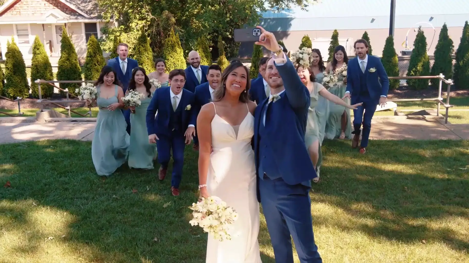 A bride and groom, Sherilyn and Alex, take a selfie as their wedding party joyfully approaches from behind. The group is outdoors on a sunny day with a house and trees in the background. Bridesmaids wear light green dresses, and groomsmen wear blue suits.
