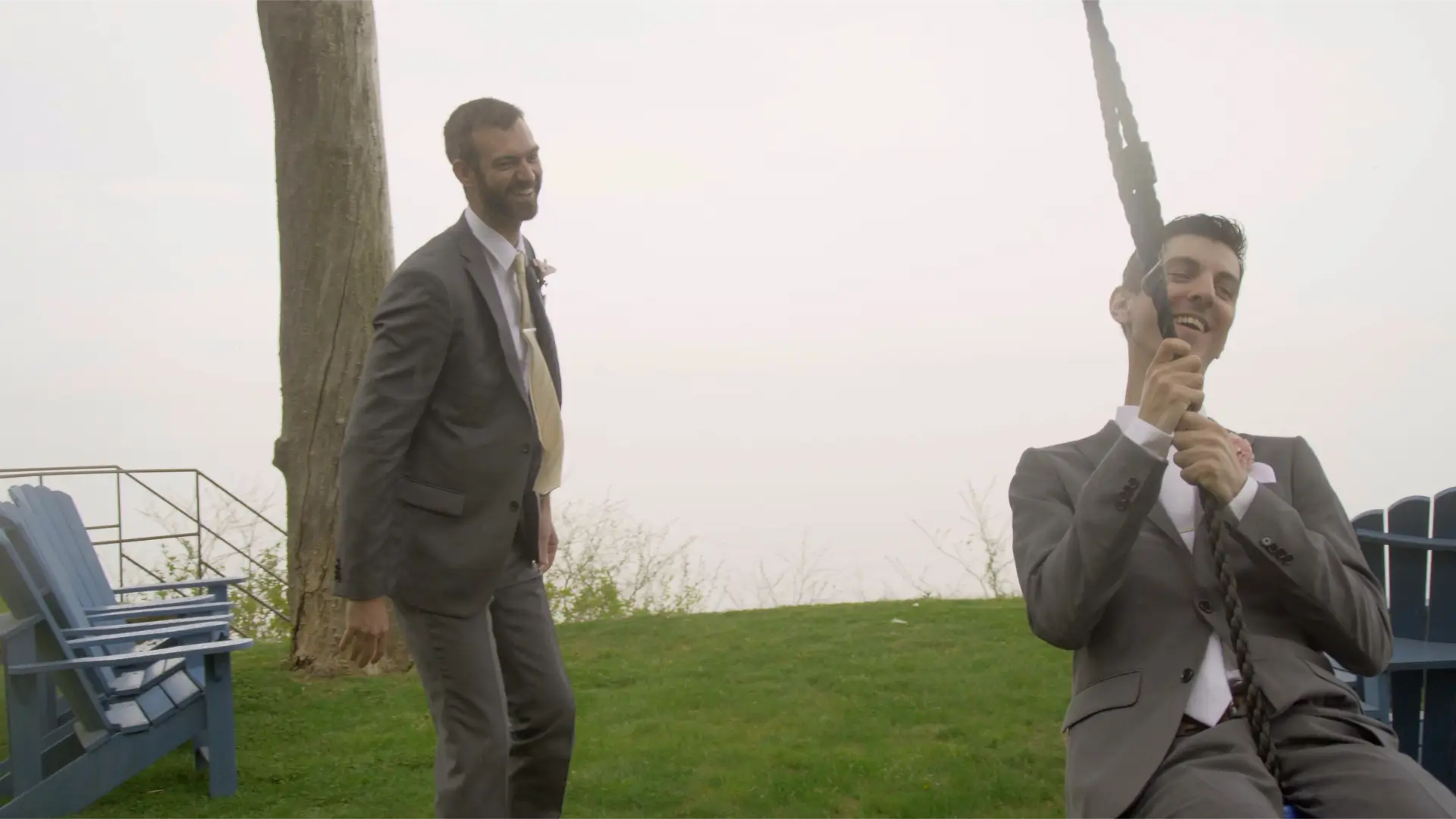 The groom and a groomsman in suits are laughing outdoors by a tree. The groom is on a swing while his groomsman stands beside him, both smiling. The background features a misty landscape with green grass and a foggy sky.