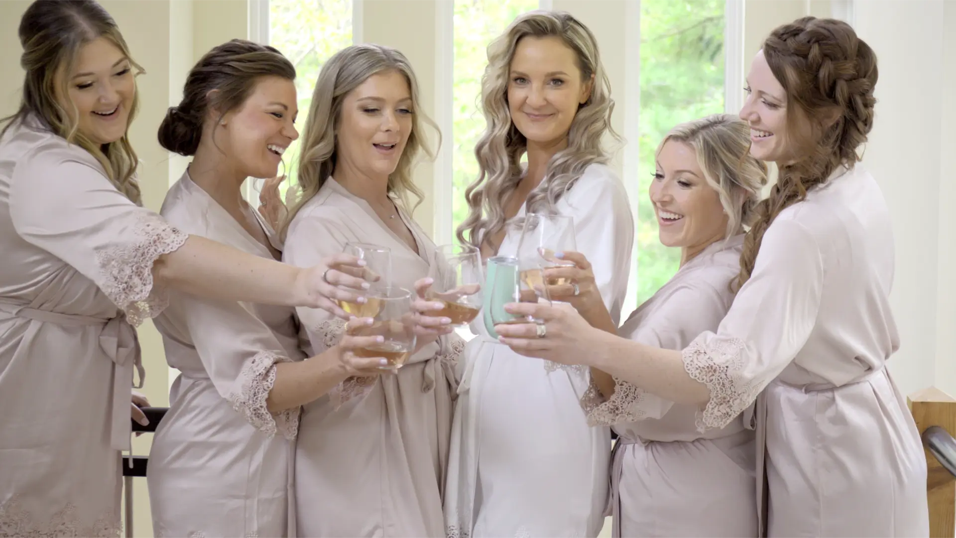 In this still from their wedding videographer, bride Jessica and her bridesmaids share a toast while getting ready at an AirBNB. They are all wearing matching robes and smiling at the camera, creating a joyous and celebratory atmosphere.