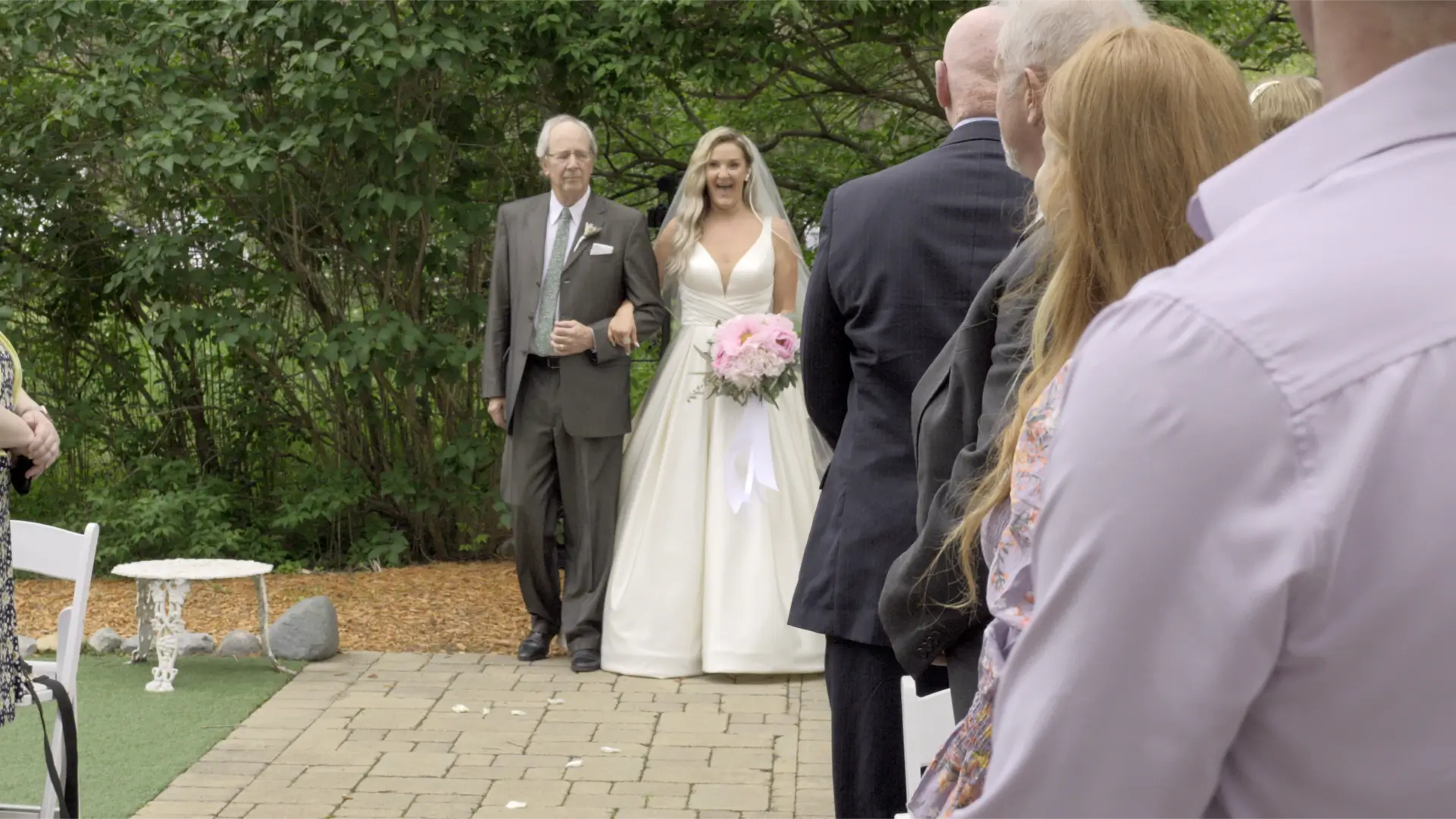 Bride Jessica, in a white gown holding pink flowers, walks down the outdoor aisle with an older gentleman. Guests dressed in formal attire watch from both sides in a lush, green garden setting, suggesting a wedding at the Blue Dress Barn.