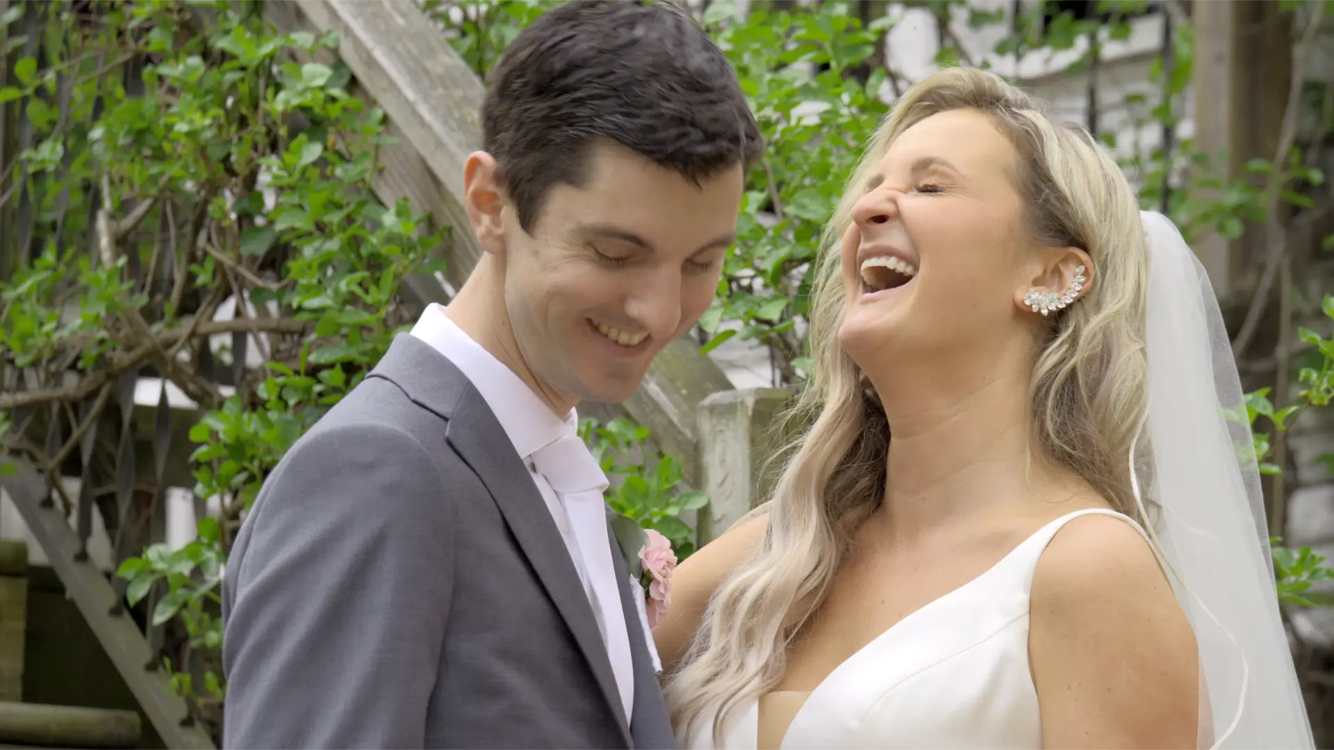 Jessica and Patrick, the bride and groom, share a joyful moment on their wedding day. Jessica laughs, wearing a white dress and veil, while Patrick, in a gray suit, smiles with his eyes closed. Greenery adds to the lively atmosphere.