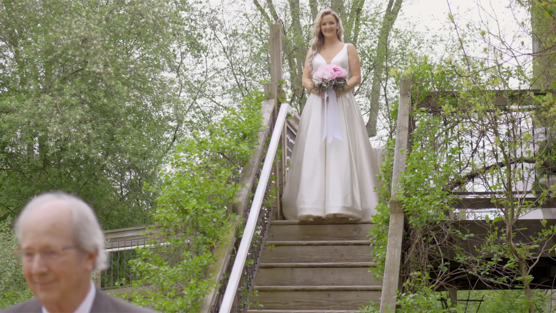 Bride Jessica stands at the top of wooden outdoor stairs holding a pink bouquet surrounded by greenery, smiling and wearing an elegant white gown. Trees and foliage frame the background, creating a lush setting. Her father in the foreground, out of focus.