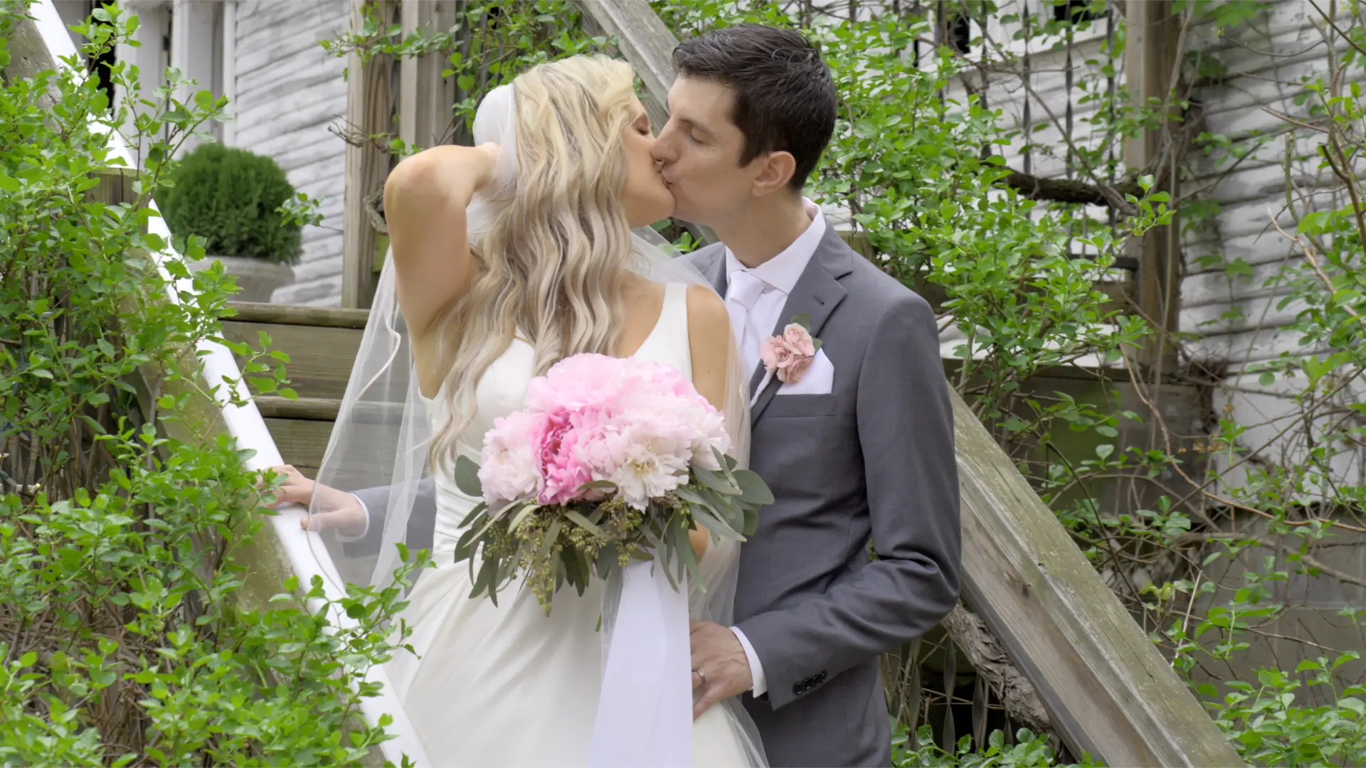 Bride Jessica and groom Patrick share a kiss outside the Blue Dress Barn, captured by their videographer. Jessica holds a bouquet of pink peonies and wears a white gown, while Patrick is in a grey suit with a pink boutonniere.