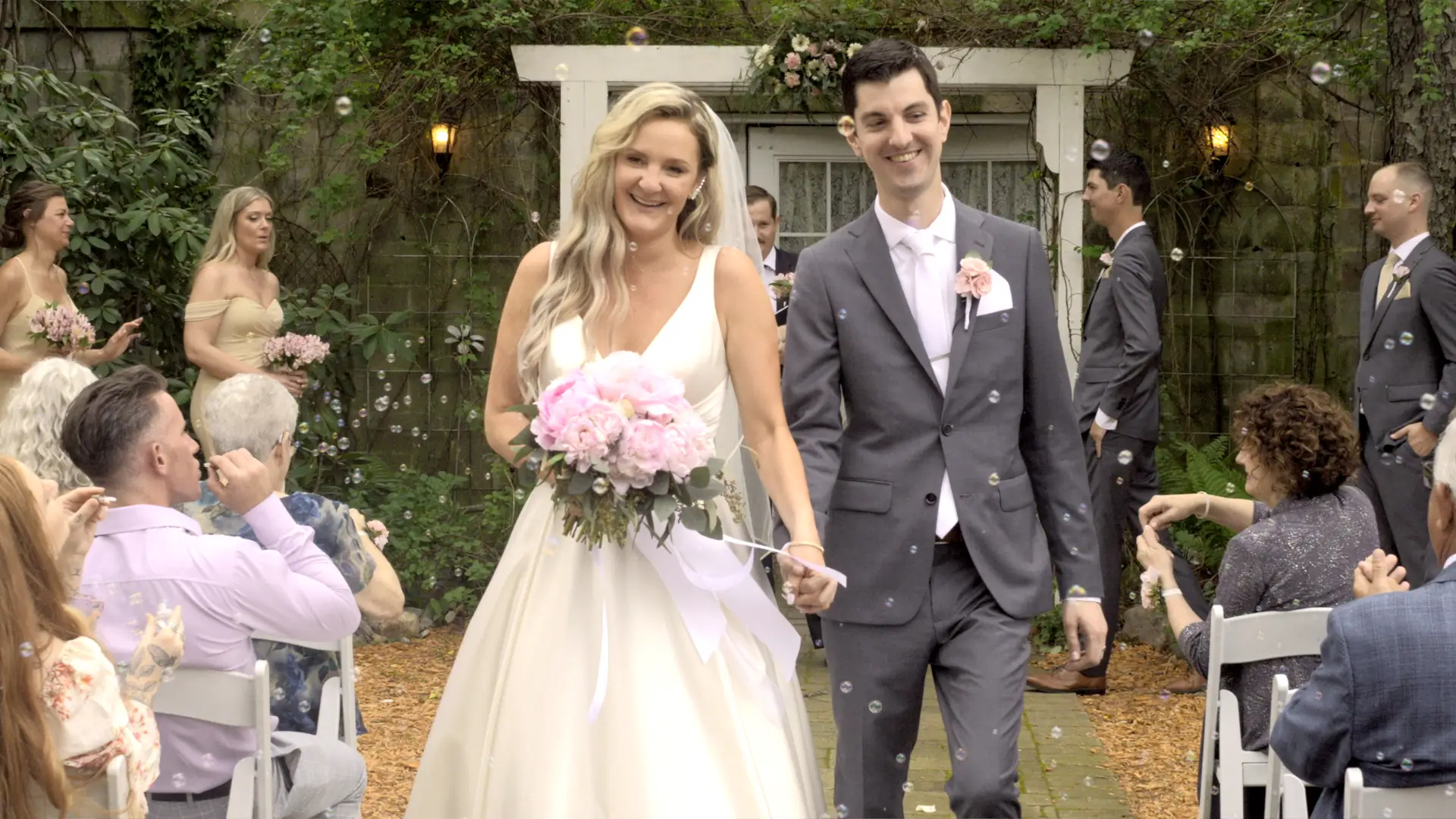 Bride and groom, Jessica and Patrick, smile and hold hands while walking down the outdoor aisle. Guests blow bubbles as they pass. Bridesmaids and groomsmen are in the background. The scene is lush and green, creating a relaxed, joyful atmosphere.