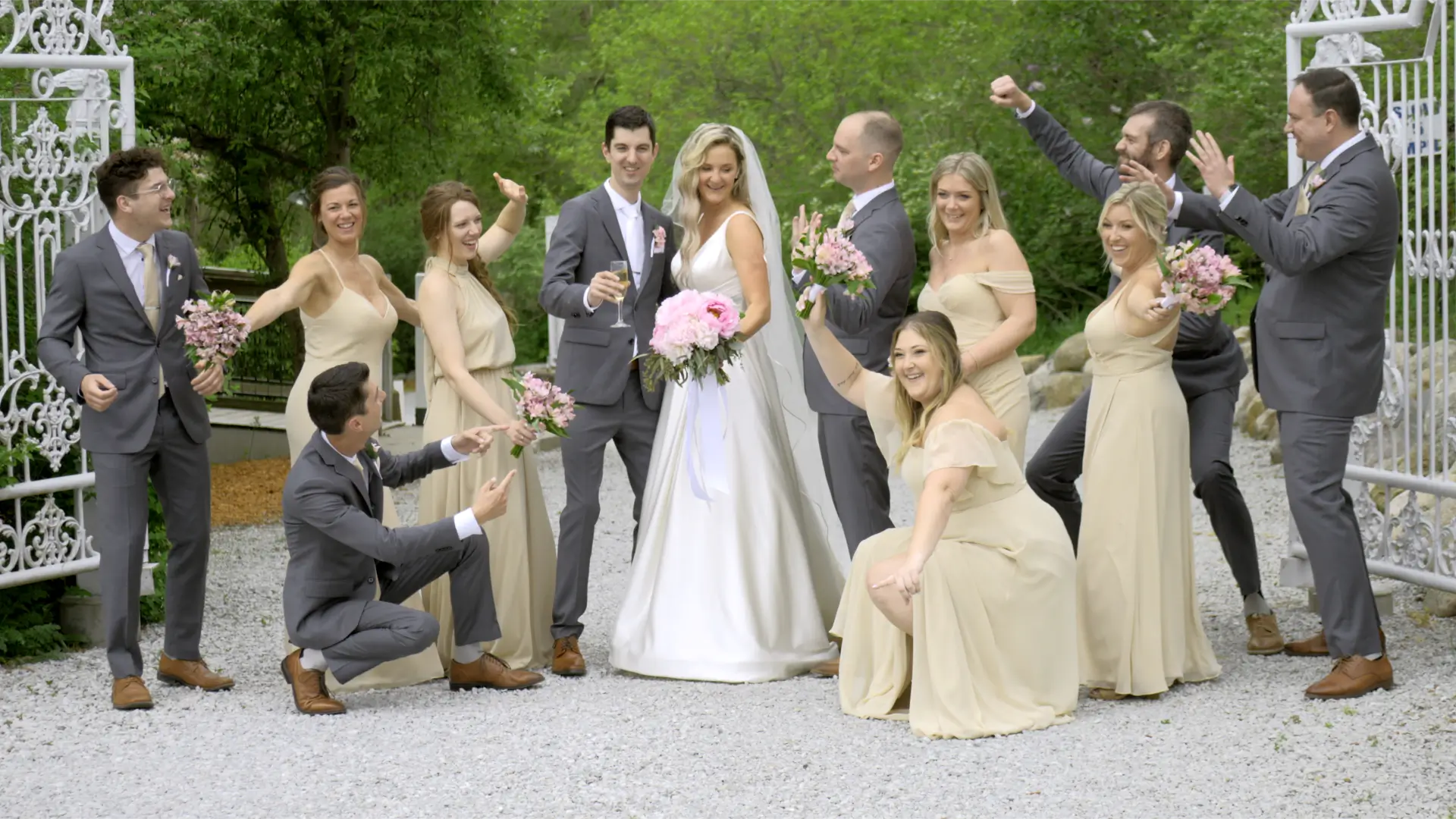 Bride Jessica and groom Patrick pose joyfully with their wedding party in a garden setting. Bridesmaids wear beige dresses and groomsmen don gray suits, all celebrating the couple's special day.