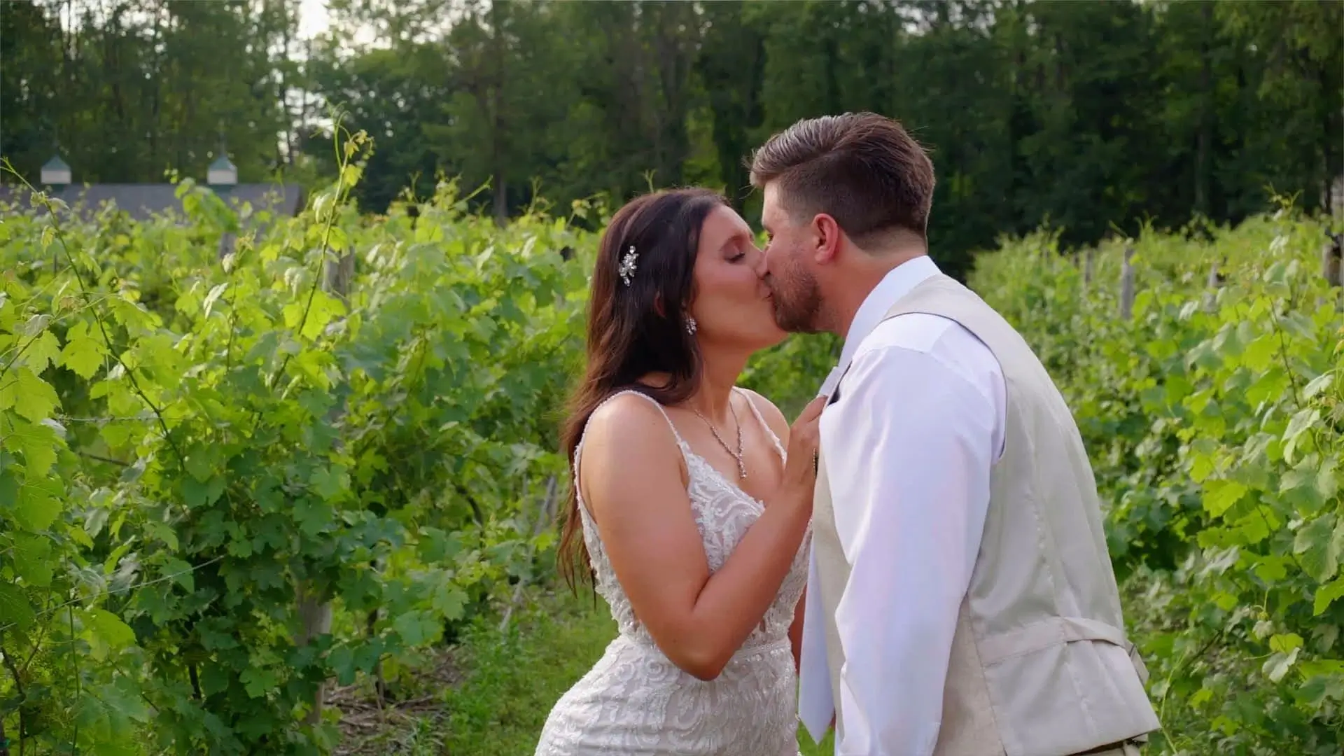 Amanda and Isaac share a kiss in a vineyard at Rooster Ridge. Amanda is in a lace wedding dress with spaghetti straps, her hand on Isaac's chest. He wears a white shirt and beige vest. Green foliage surrounds them, creating a romantic atmosphere.