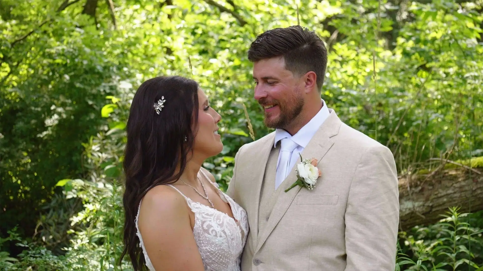 Amanda and Isaac stand facing each other in a lush green setting at Rooster Ridge Vineyard, New Era, Michigan. Amanda wears a lace wedding dress and a hair accessory, while Isaac is in a light beige suit with a white tie and boutonniere. Both are smiling.