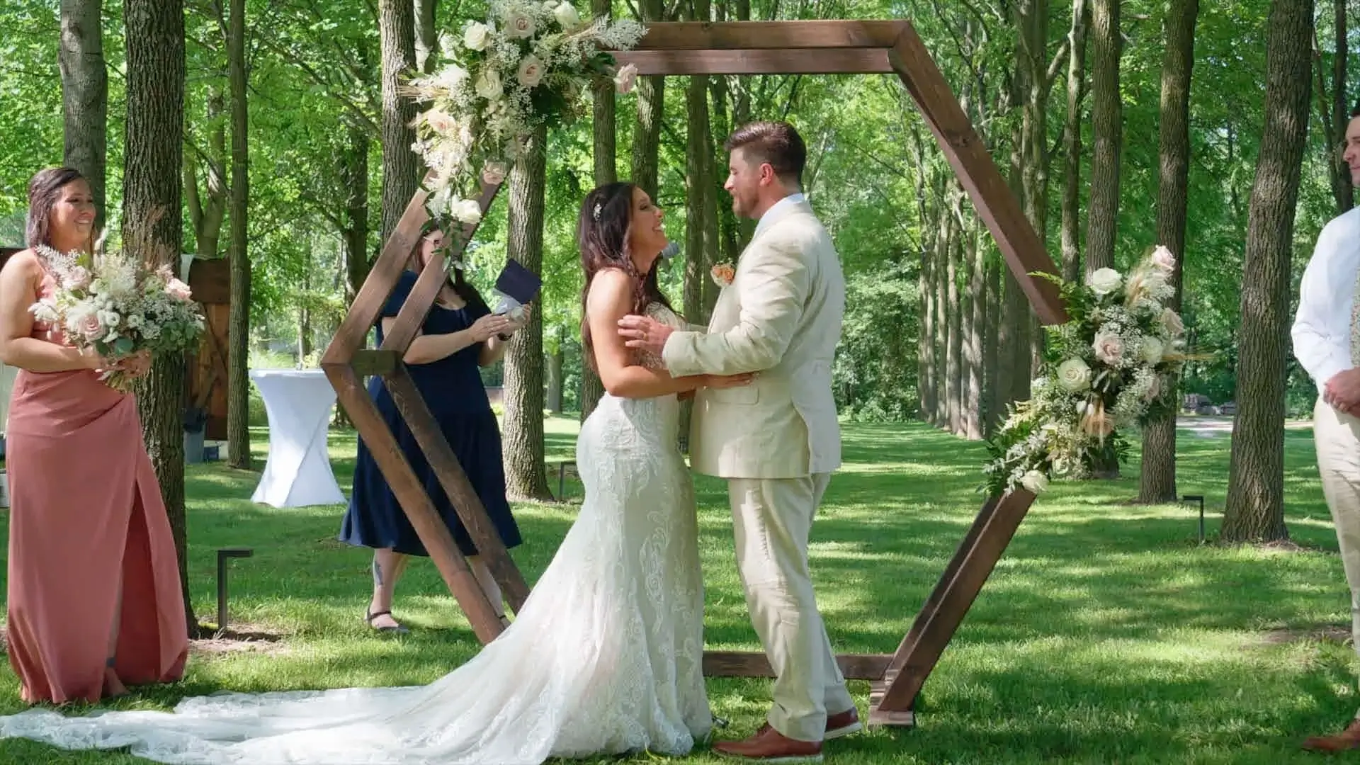 Amanda & Isaac stand under a hexagonal wooden arbor adorned with flowers during their wedding at Rooster Ridge Vineyard. The bride wears a lace gown, and the groom is in a light beige suit. 