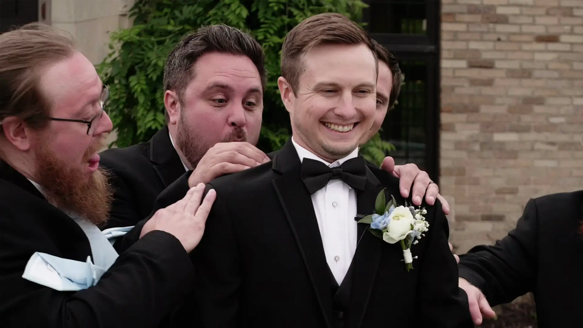 A group of smiling groomsmen in tuxedos, playfully adjusting the groom's bow tie before the wedding ceremony at Venue3Two in Grand Rapids, Michigan. The groom has a boutonnière with white and blue flowers.
