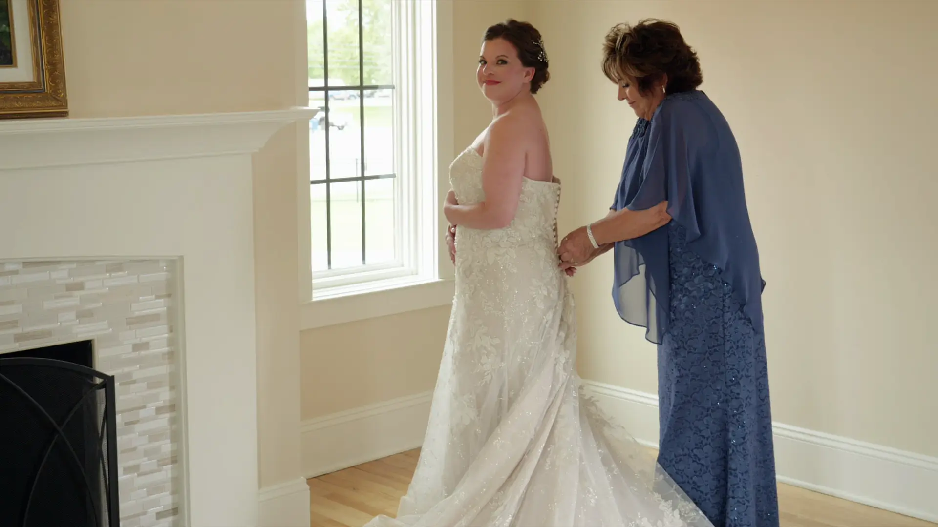 A bride, Leah, stands in a lovely bridal gown with intricate lace details beside a woman helping to fasten the dress. They are in a well-lit room with a large window and a fireplace, preparing for a wedding at Venue3Two in Grand Rapids, Michigan.