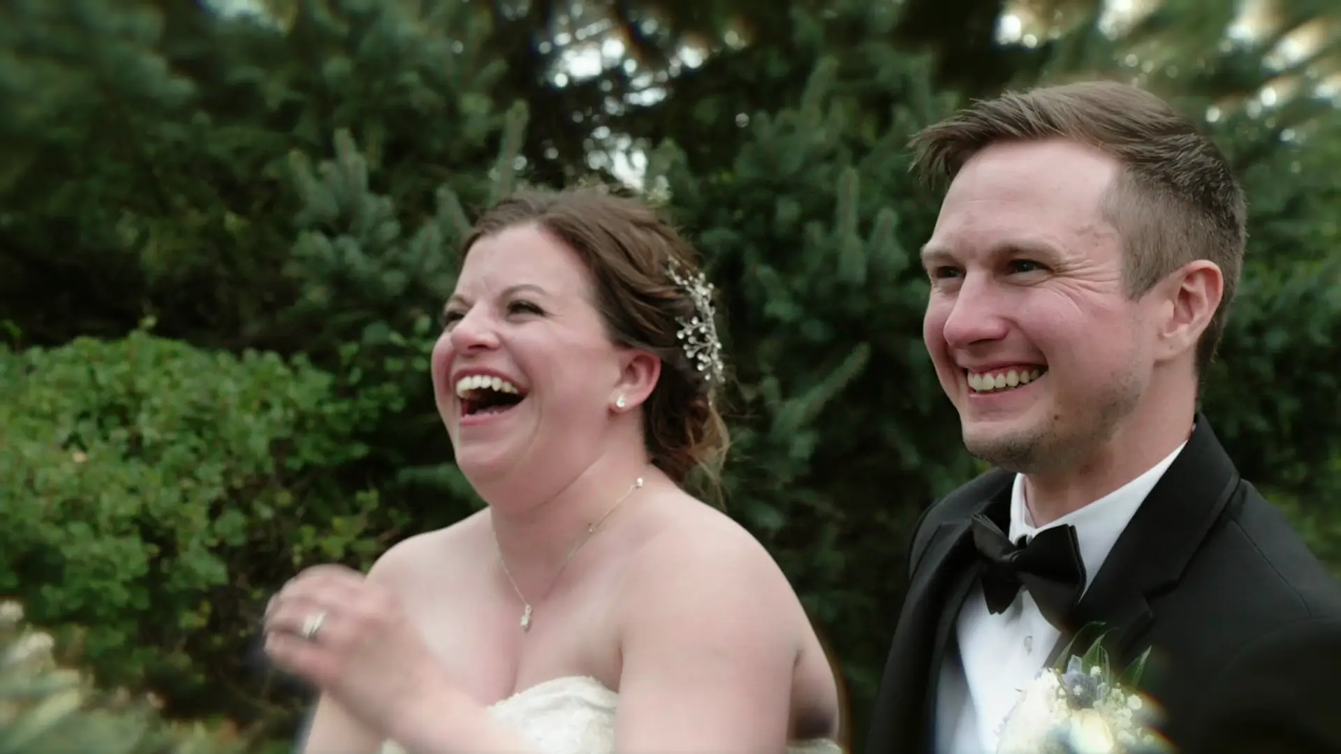 A bride and groom, Leah and Eric, are caught in a candid moment of laughter in Grand Rapids, Michigan. The bride wears a strapless gown with floral hair accessories, and the groom is in a tuxedo with a boutonniere, set against a backdrop of greenery.