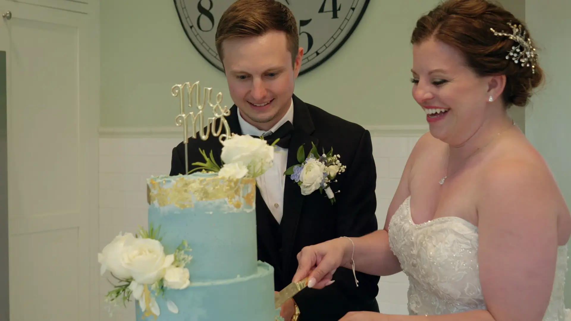 Leah and Eric cutting a blue and gold wedding cake with floral decorations at Venue3Two in Grand Rapids, Michigan. Leah is in a white strapless gown with an embellished hairstyle, while Eric wears a black tuxedo with a boutonniere.