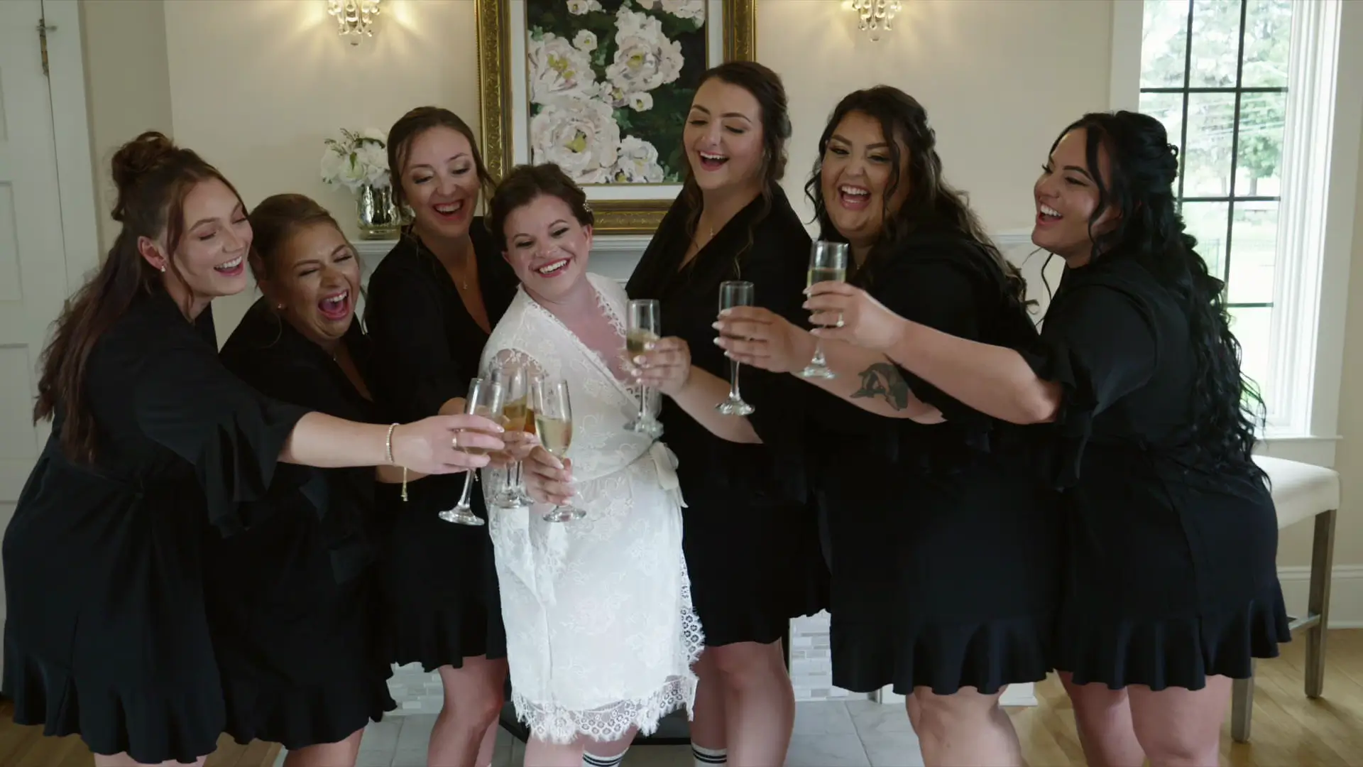 Bride Leah with bridesmaids in black robes, all smiling and raising champagne glasses, standing indoors at Venue3Two in Grand Rapids, Michigan. A floral painting is visible in the background.