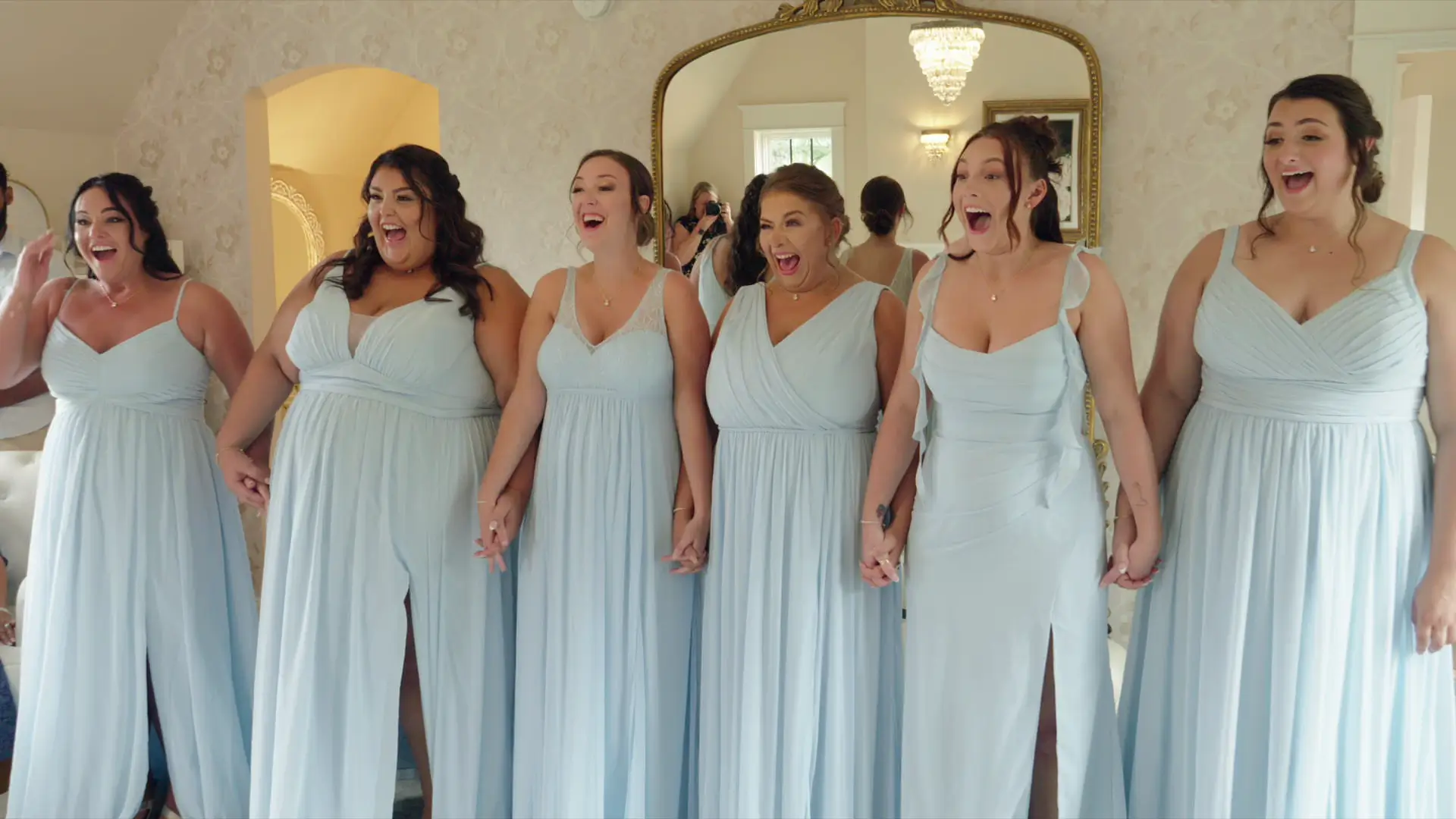 Six bridesmaids in light blue dresses hold hands and express excitement as they see bride Leah during their bridesmaid first look.