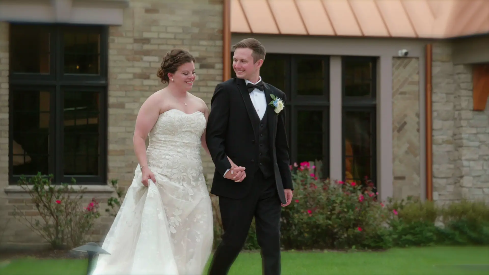 A bride and groom, Leah and Eric, walk hand in hand outside Venue3Two in Grand Rapids, Michigan, against a backdrop of blooming roses and classic architecture, exuding elegance and happiness on their wedding day.