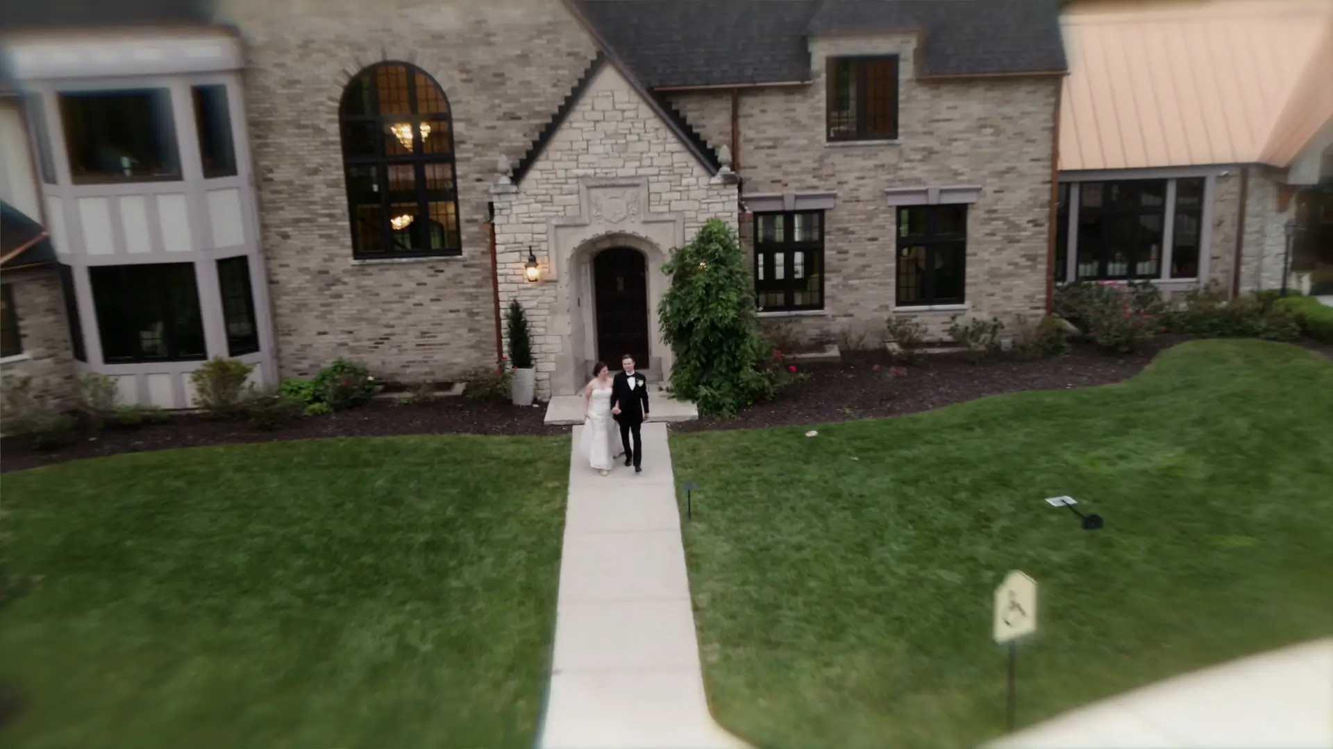 
Bride and groom walk hand in hand on a path in front of charming Venue3Two surrounded by lush Michigan greenery, captured from high above by drone video on their elegant wedding day.