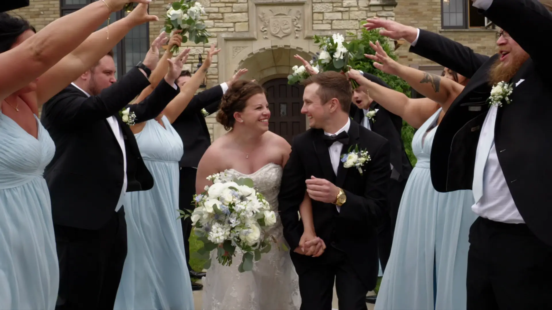 Bride Leah and groom Eric walk joyfully through an arch of raised arms and bouquets, surrounded by their wedding party in elegant attire at Venue3Two, Grand Rapids, Michigan. Leah holds a bouquet of white flowers, and both are smiling at each other.