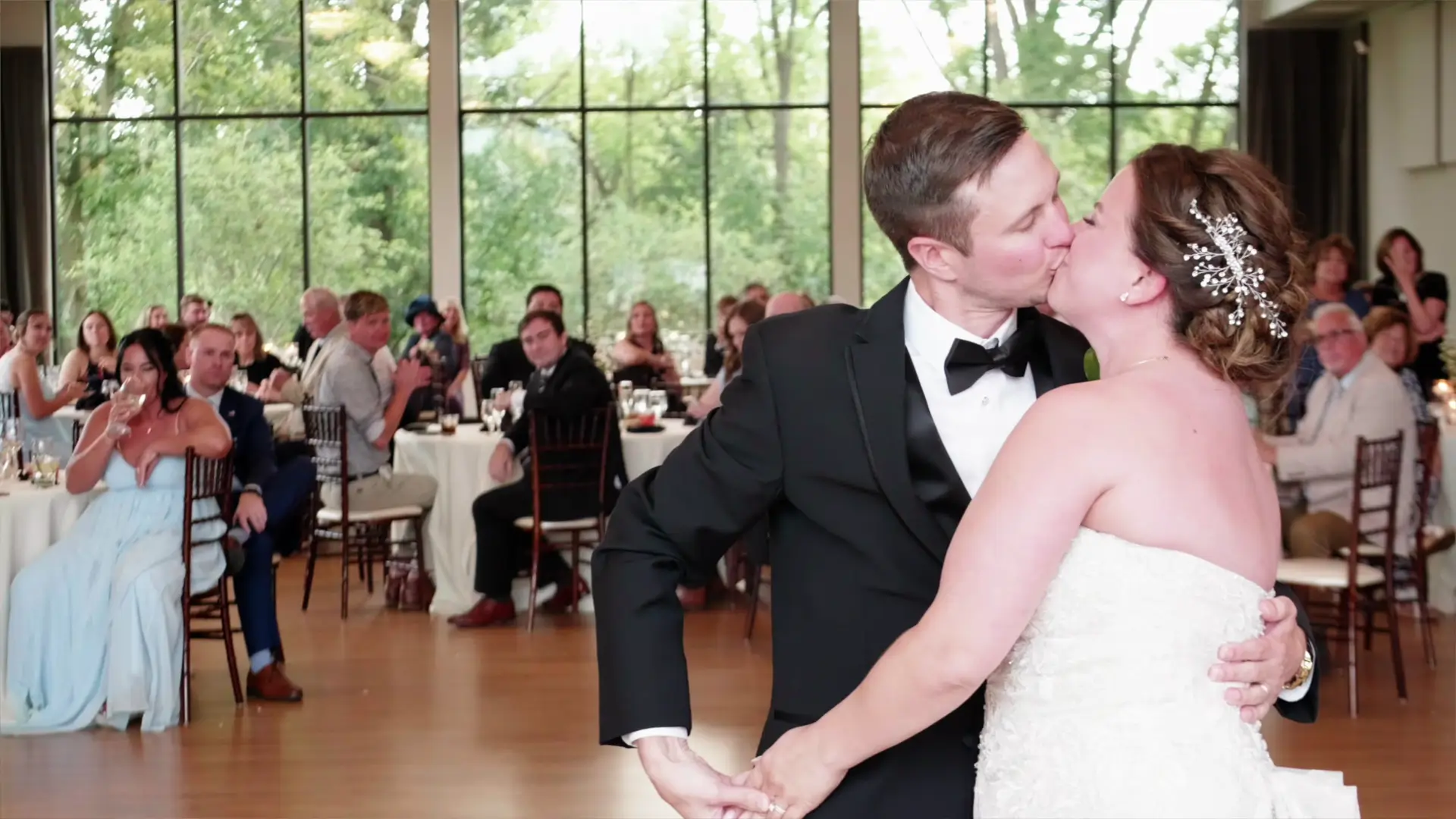 In this wedding videographer's snapshot, Leah and Eric share a kiss during their first dance at Venue3Two in Grand Rapids, Michigan, surrounded by guests and vibrant greenery through the large windows.