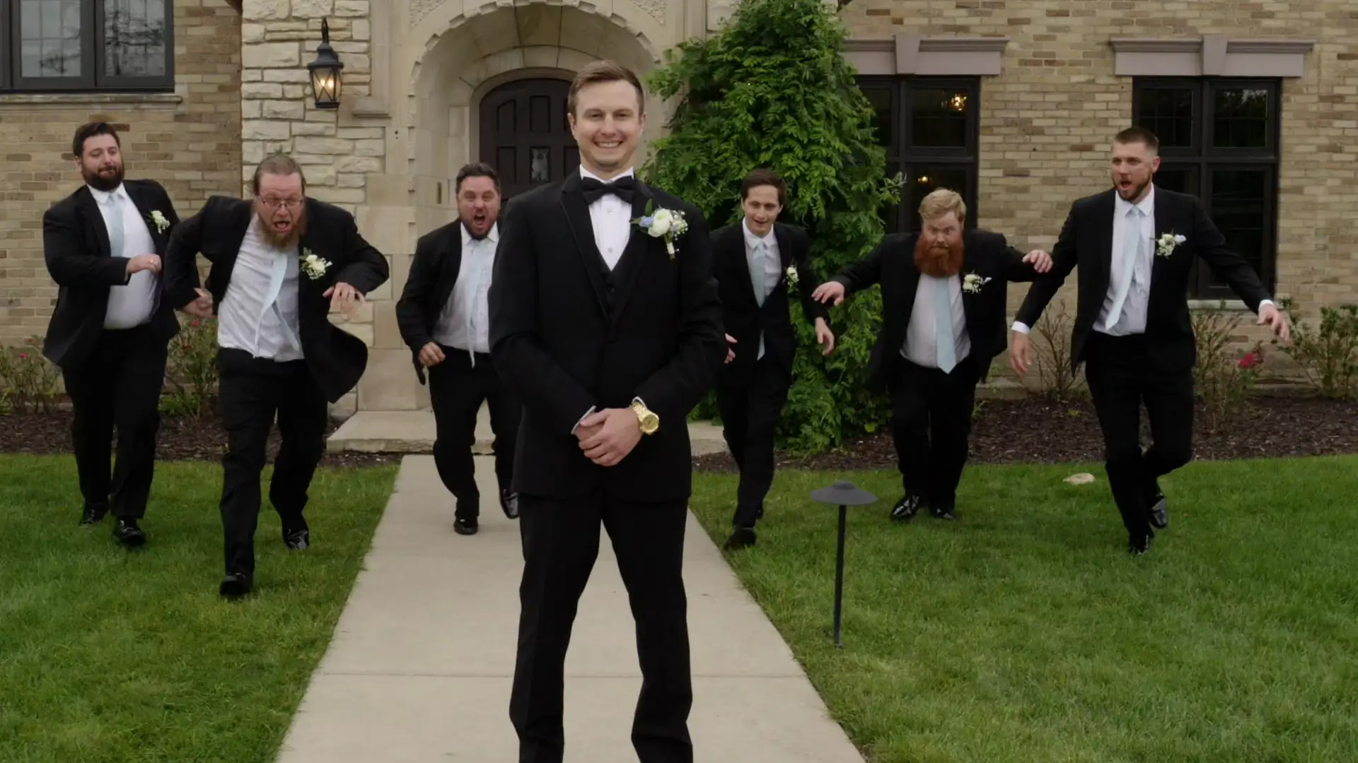Groom Eric poses confidently in front of groomsmen playfully running behind him at Venue3Two in Grand Rapids, Michigan. The men are dressed in formal black suits, adding to the elegant wedding theme.