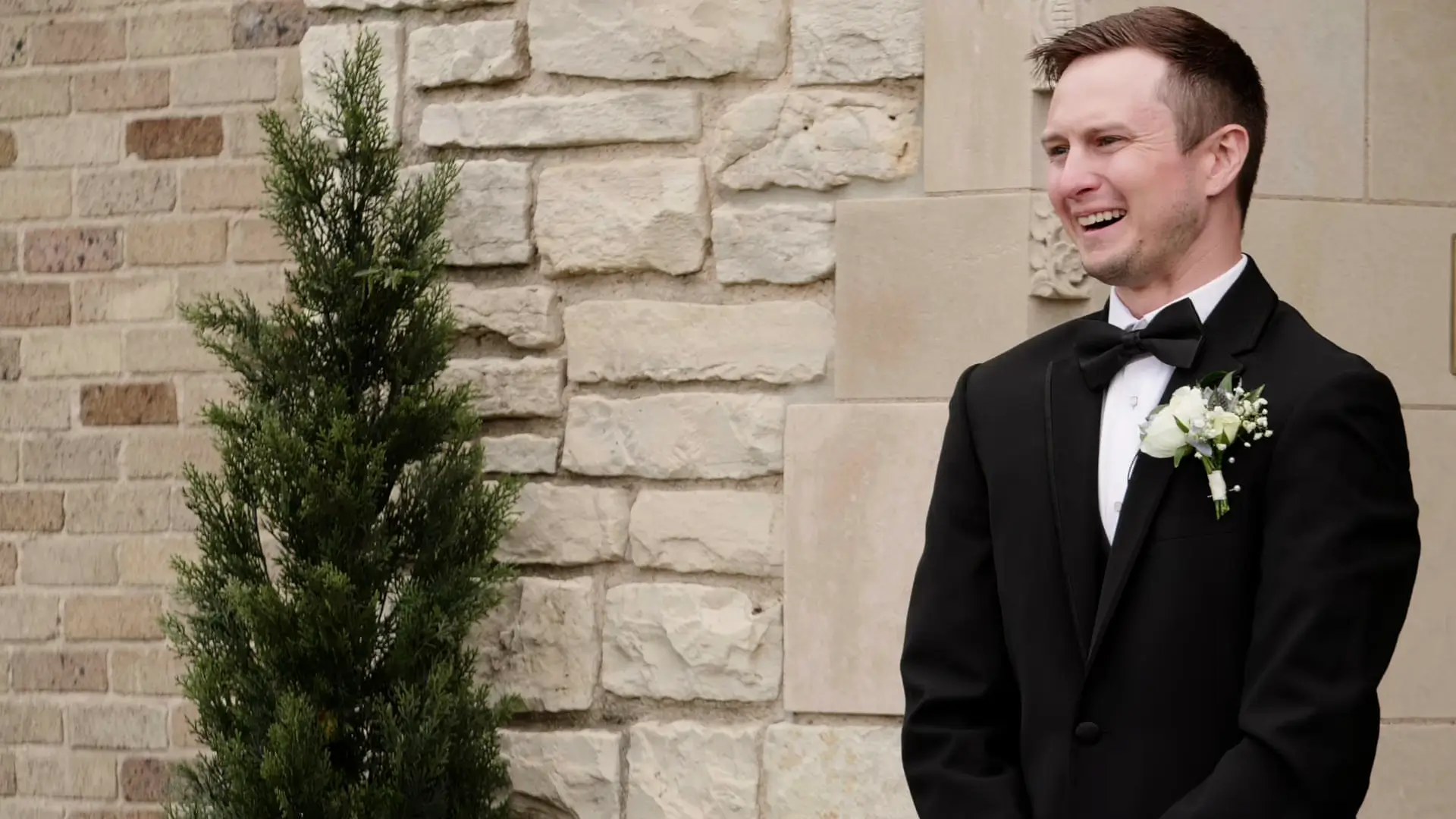 Groom Eric wears a black tuxedo with a boutonniere, and smiles with tears in his eyes as he beholds his bride during this video snapshot of their first look. He is in front of a stone wall at Venue3Two in Grand Rapids, Michigan.