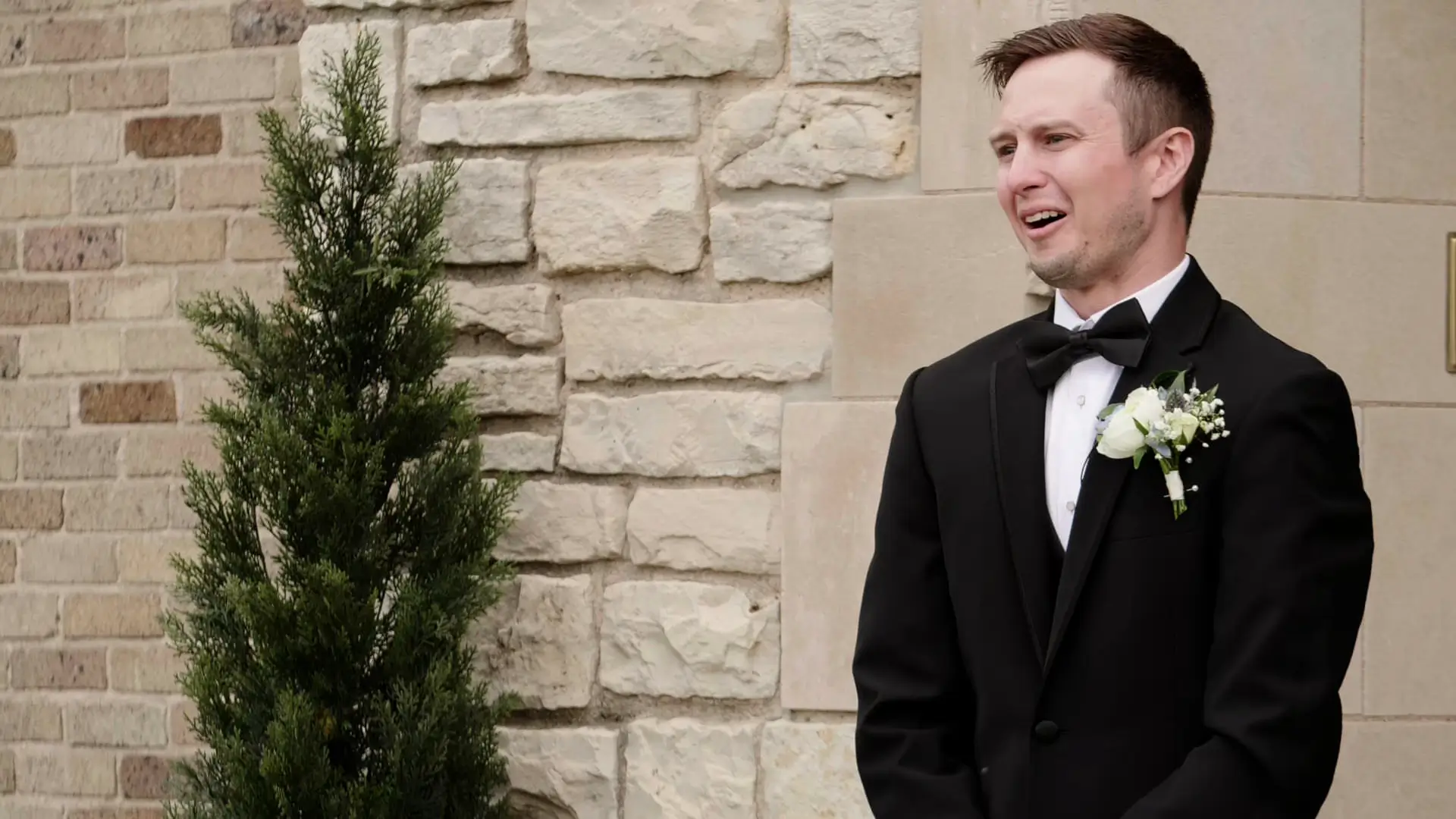 Groom in a black tuxedo stands against a stone wall, looking emotional as he beholds his bride during their first look. A small evergreen tree adds a touch of Michigan greenery to the scene.